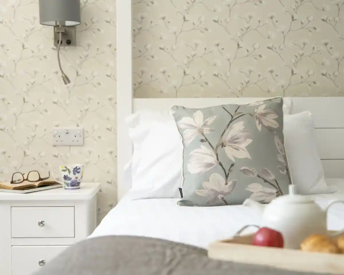 A cozy bedroom with a white bed featuring a decorative floral pillow. A bedside table holds a cup, glasses, and a small wooden tray with an apple and pastries. The wall has a light floral wallpaper.