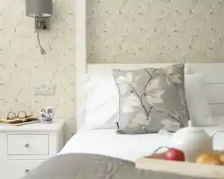 A cozy bedroom with a white bed featuring a decorative floral pillow. A bedside table holds a cup, glasses, and a small wooden tray with an apple and pastries. The wall has a light floral wallpaper.