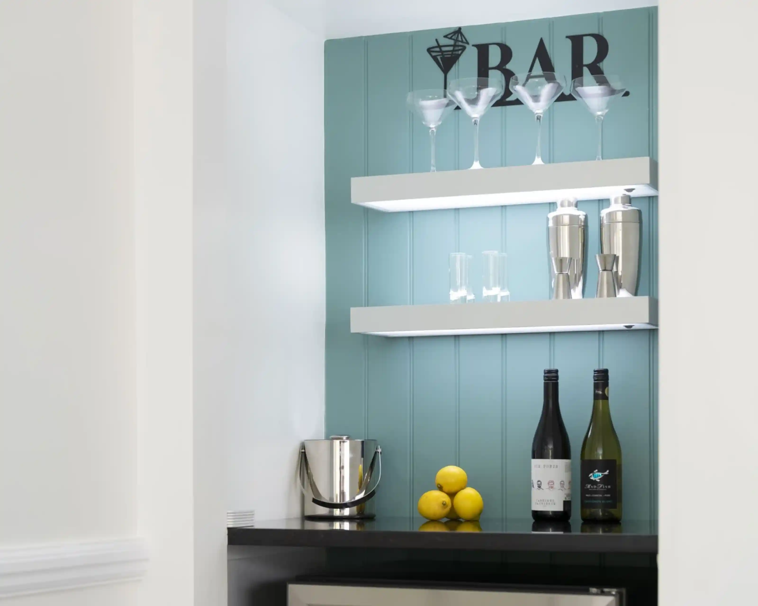 A stylish home bar area with light blue wood paneling. Two white shelves display martini glasses and cocktail shakers. Below, there are two bottles of wine and a silver ice bucket with three lemons next to it. A small refrigerator is also visible.
