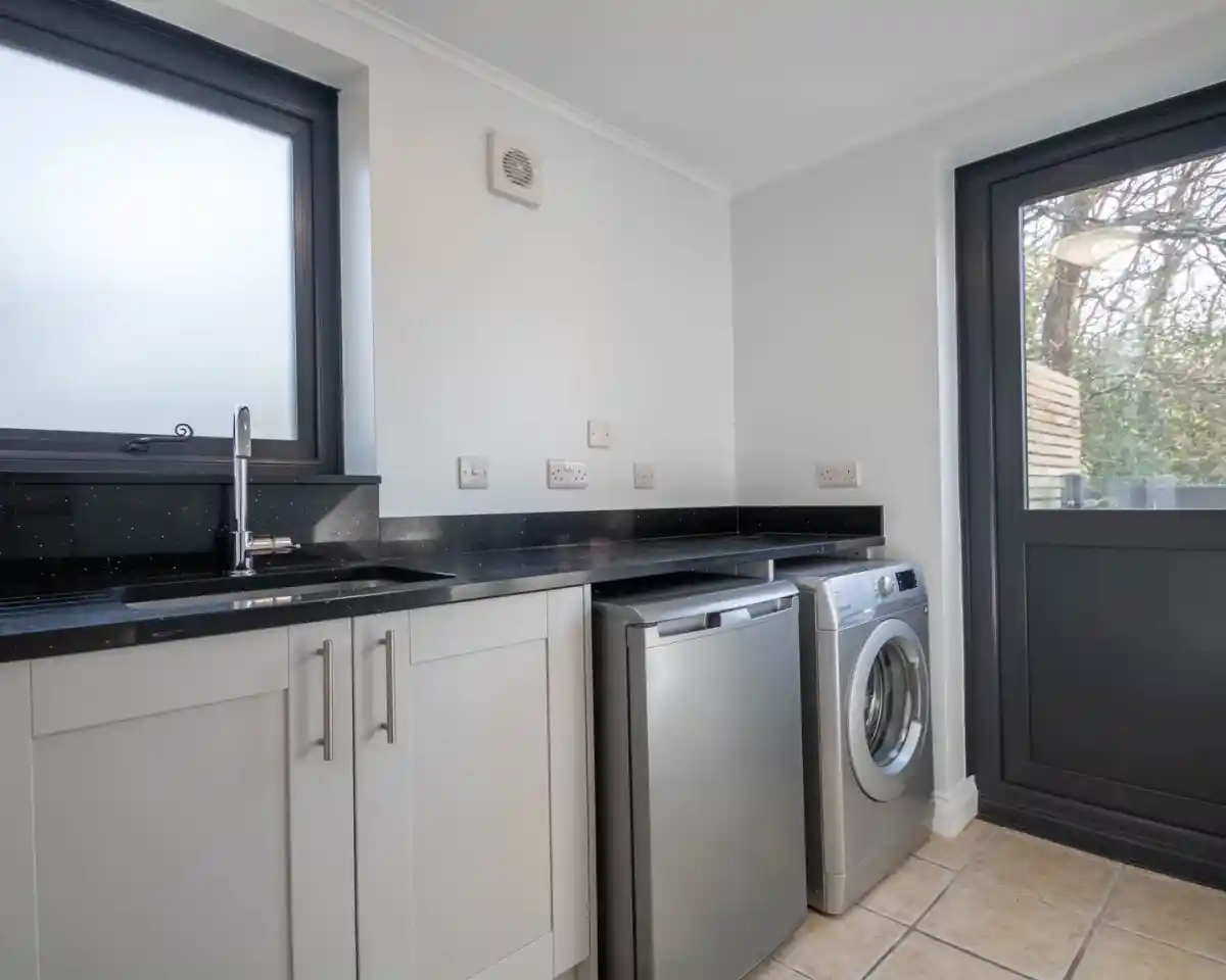 A modern laundry room featuring a stainless steel washer and dryer, a black countertop, and white cabinetry. Natural light streams through a window, illuminating the space.