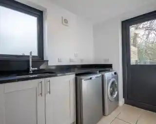 A modern laundry room featuring a stainless steel washer and dryer, a black countertop, and white cabinetry. Natural light streams through a window, illuminating the space.