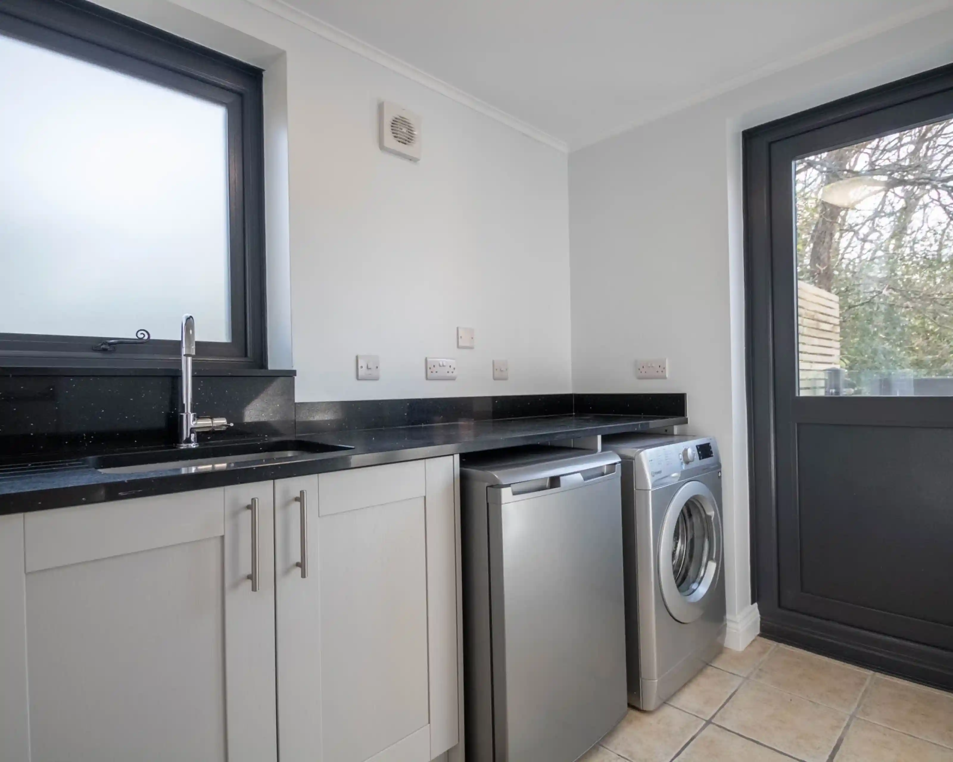 A modern laundry room featuring a stainless steel washer and dryer, a black countertop, and white cabinetry. Natural light streams through a window, illuminating the space.