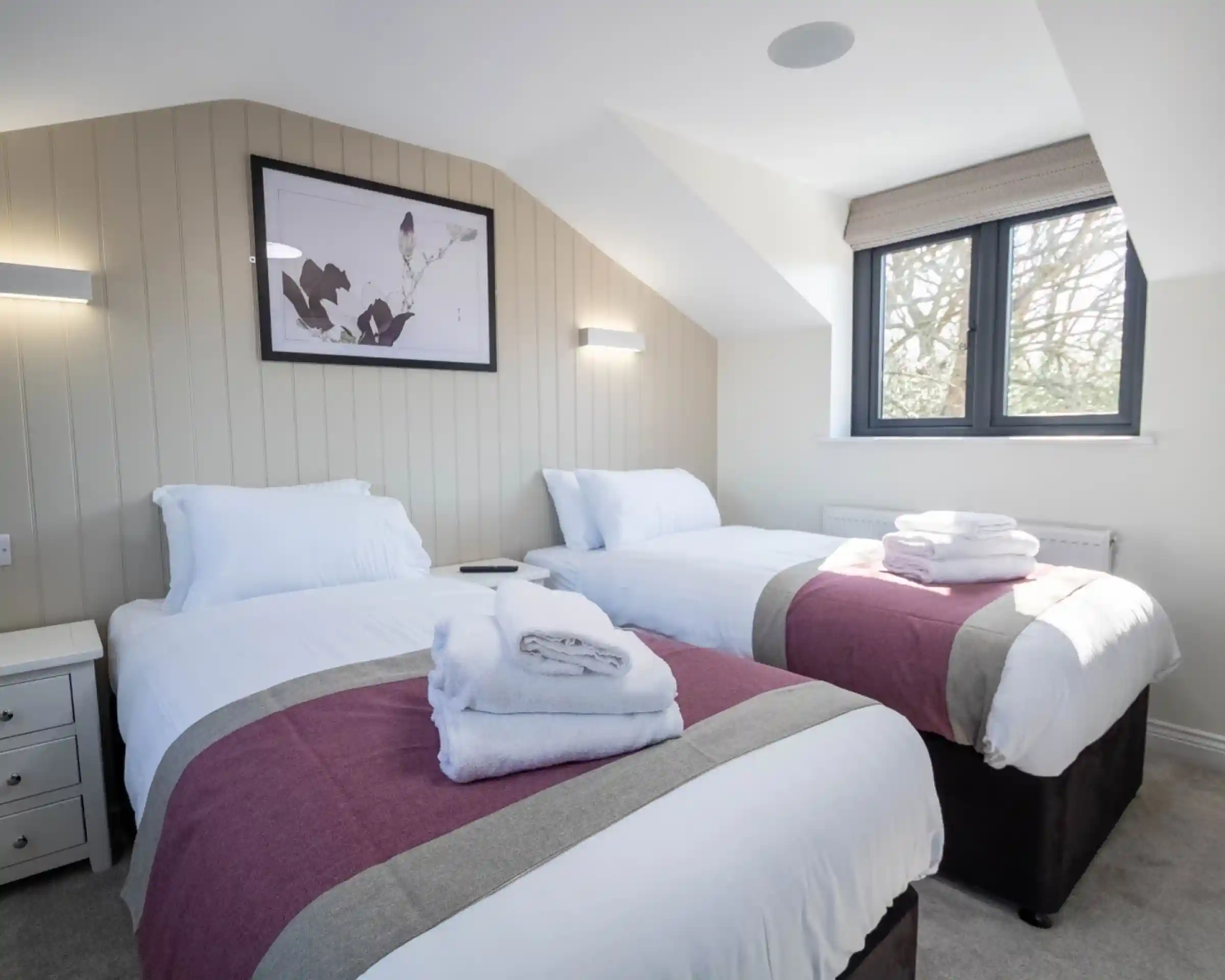 A cozy guestroom featuring two single beds with white linens and gray throws. A small nightstand is beside one bed, and a large window allows natural light to fill the space. Wall lights provide additional illumination, and a framed artwork hangs on the wall.