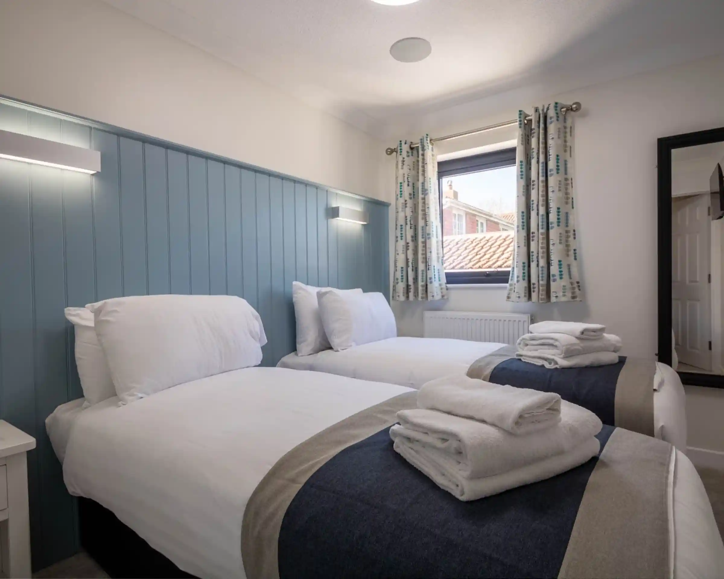 A cozy bedroom with two twin beds dressed in white linens and neatly folded towels. Light blue paneling and curtains with a patterned design complement the space. A window allows natural light, and a full-length mirror is visible in the corner.