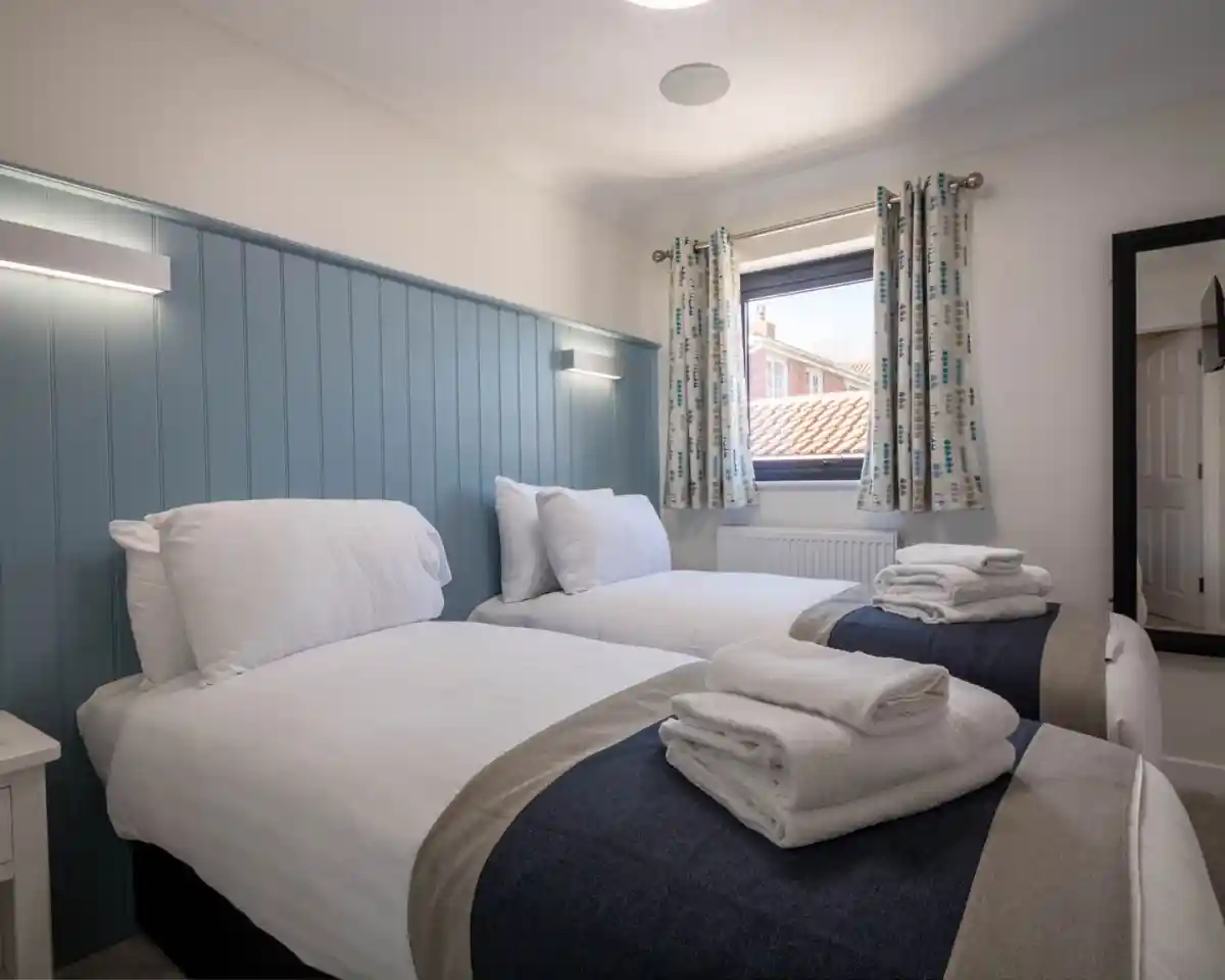 A cozy bedroom with two twin beds dressed in white linens and neatly folded towels. Light blue paneling and curtains with a patterned design complement the space. A window allows natural light, and a full-length mirror is visible in the corner.