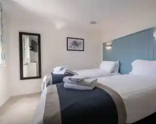 A well-lit room with two twin beds, each topped with white linens and gray towels. A blue accent wall and a mirror enhance the space, which features light-colored carpet and a picture hanging on the wall.