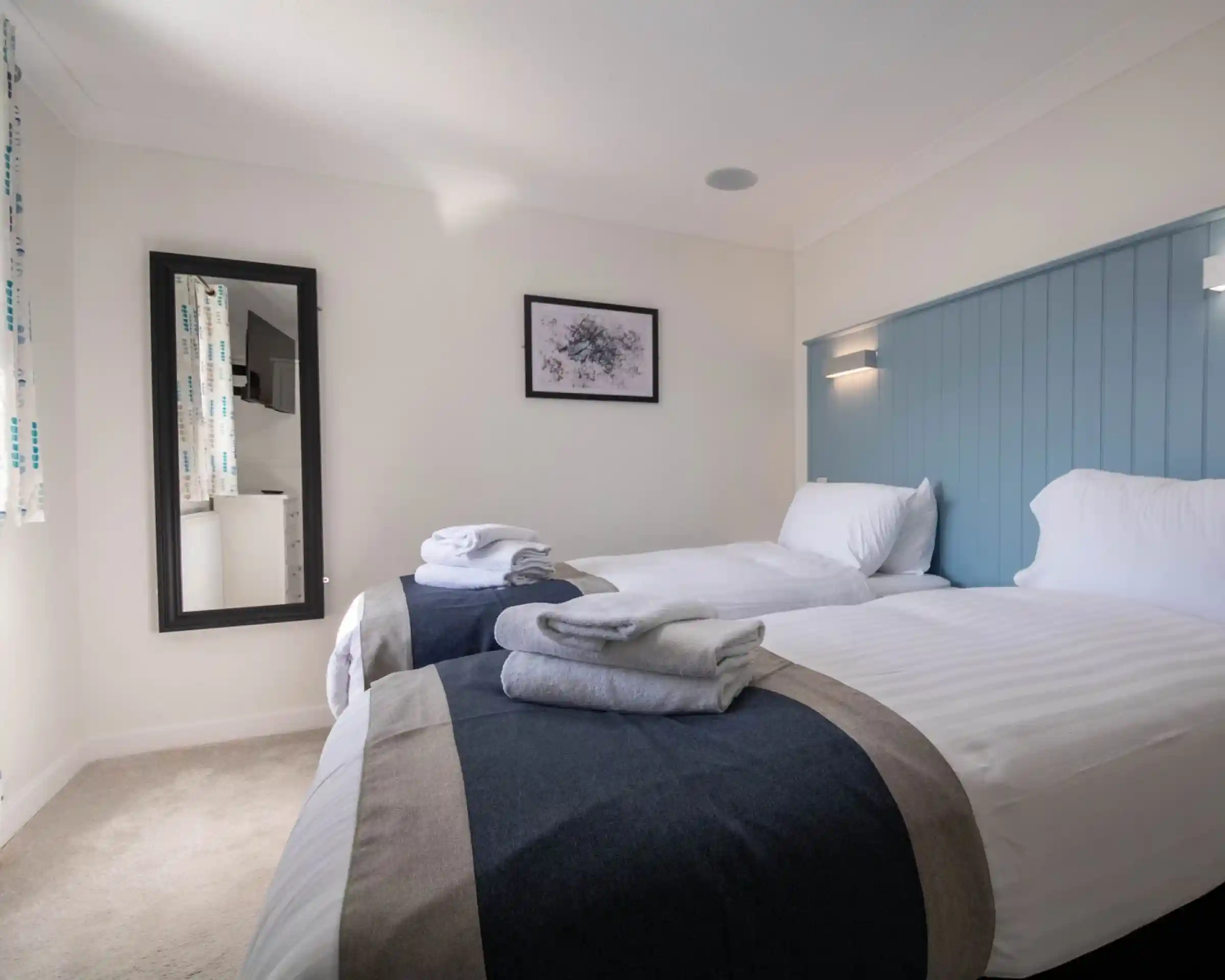 A well-lit room with two twin beds, each topped with white linens and gray towels. A blue accent wall and a mirror enhance the space, which features light-colored carpet and a picture hanging on the wall.