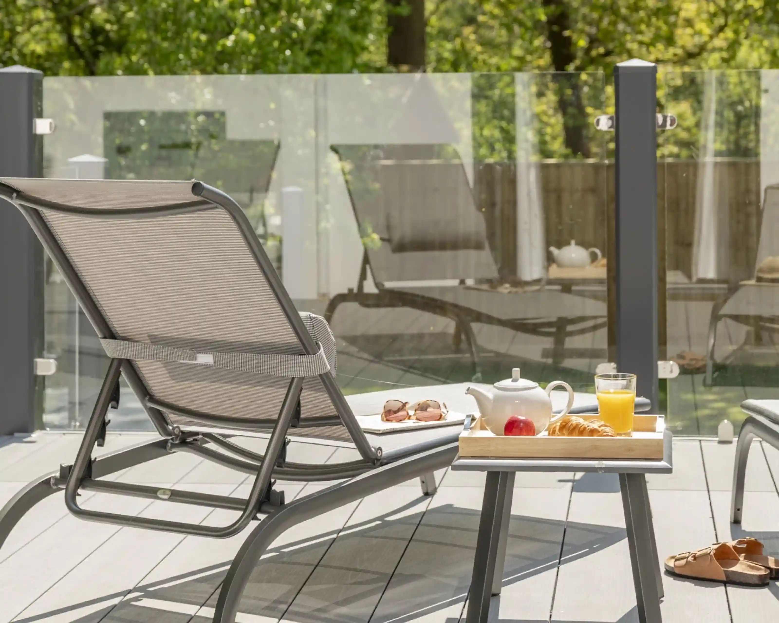A patio setting features a lounge chair, a tray with a teapot, a glass of juice, and a croissant, alongside a sunny outdoor scene with greenery in the background.