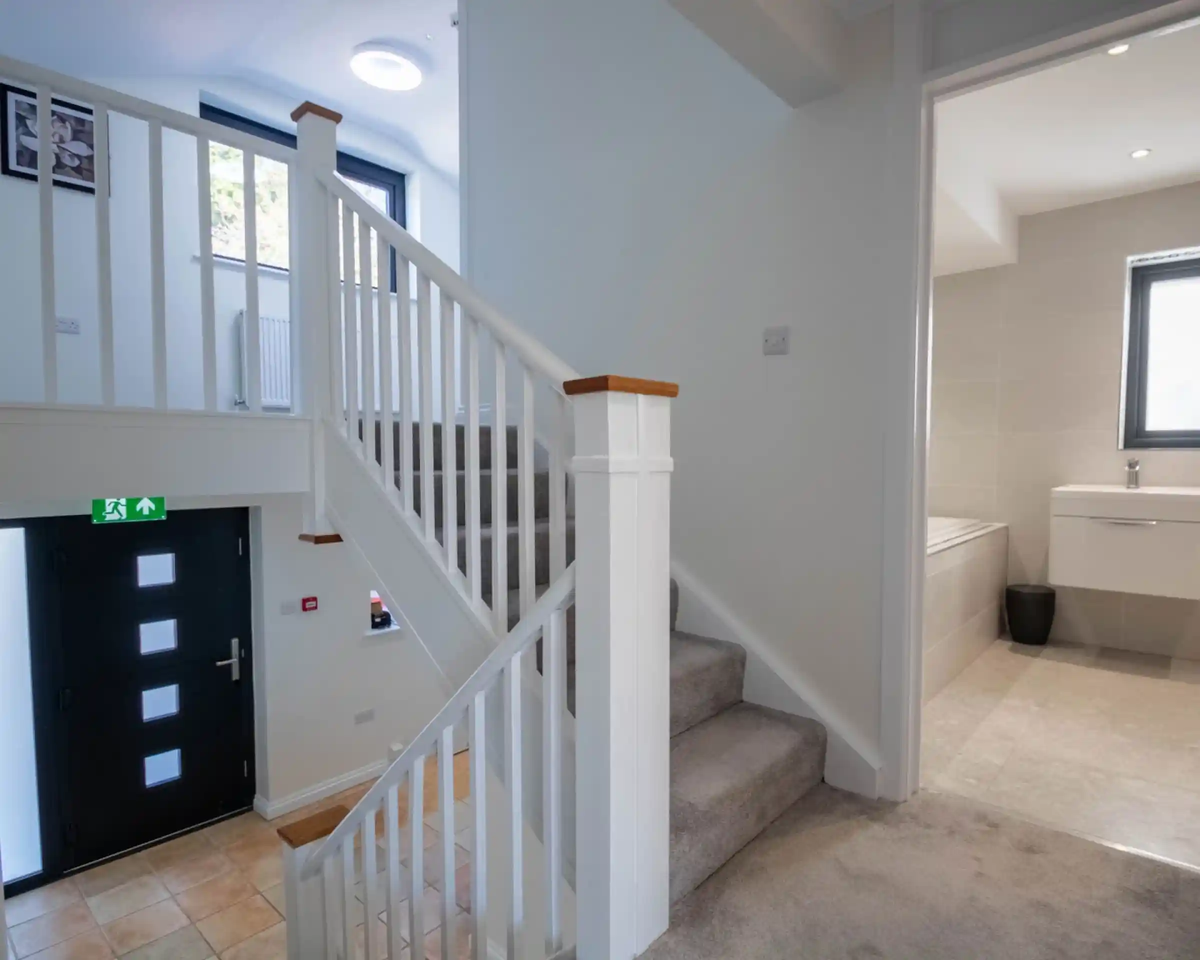 A bright interior view of a staircase leading to an upper level, with a white railing and carpeted steps. The hallway features a doorway to a bathroom on the right, and a modern front door visible on the left.