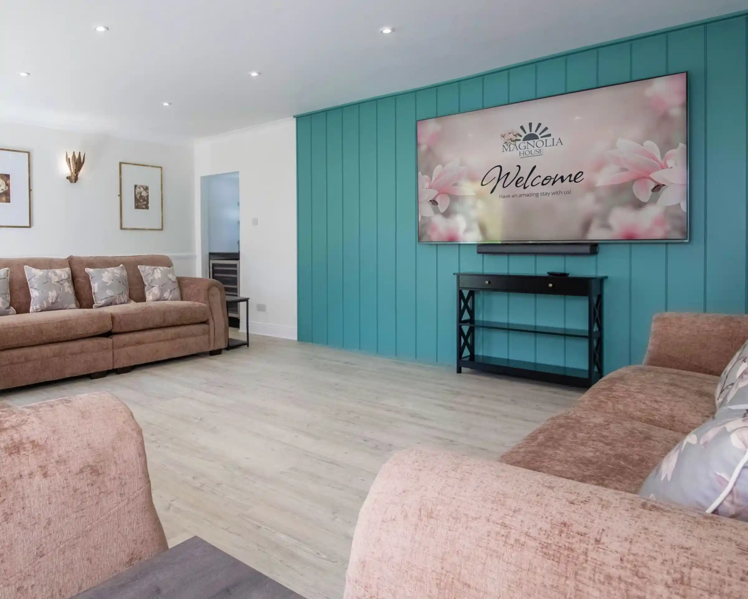A modern living room featuring two brown sofas with floral pillows, a large wall-mounted television displaying a "Welcome" message, and a stylish console table. The walls are painted light turquoise, complemented by wooden flooring.