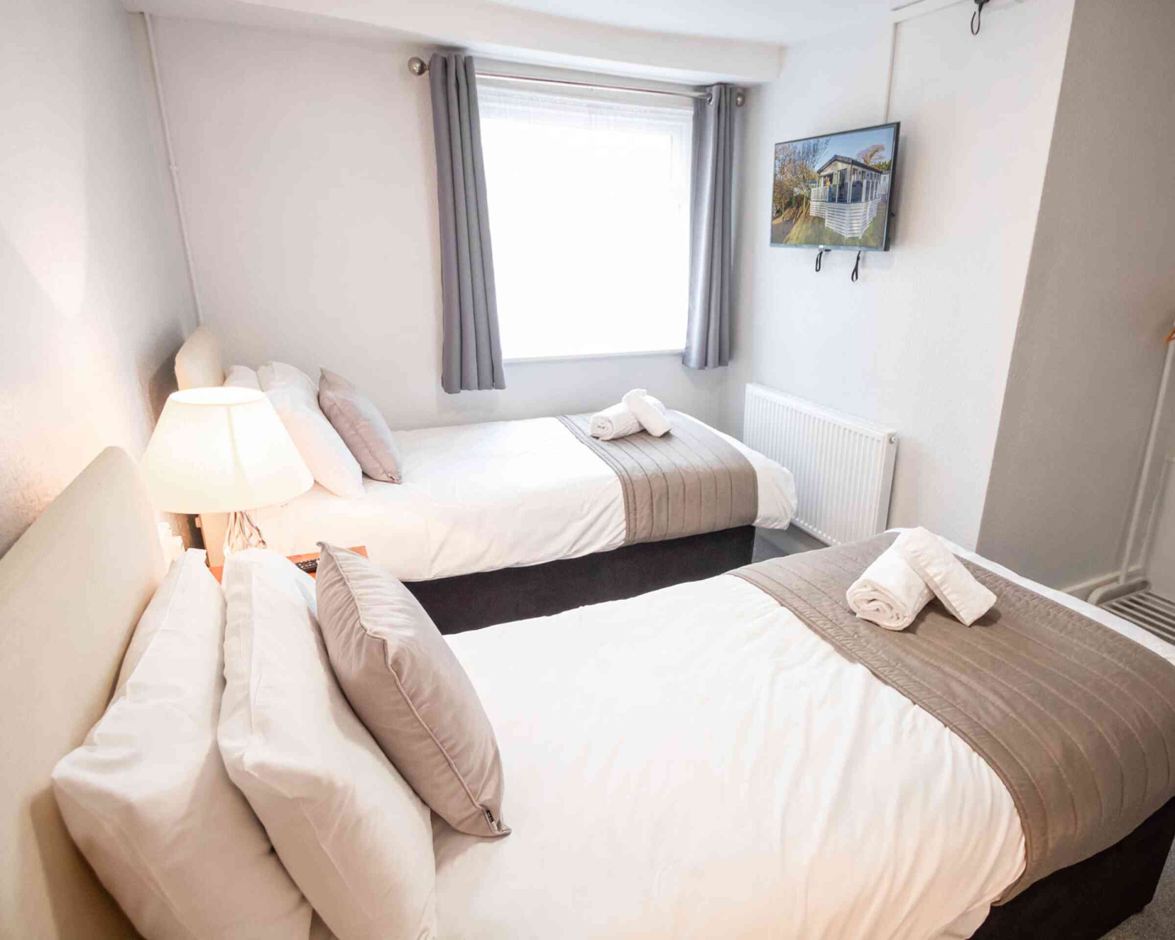 A cozy bedroom featuring two twin beds with white linens and gray accents. A lamp illuminates the room, and a flat-screen TV is mounted on the wall opposite the beds. Soft gray curtains frame the window.