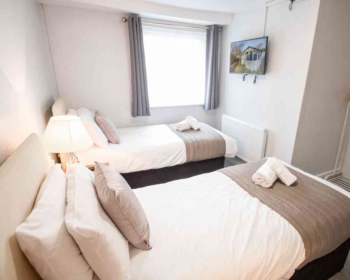 A cozy bedroom featuring two twin beds with white linens and gray accents. A lamp illuminates the room, and a flat-screen TV is mounted on the wall opposite the beds. Soft gray curtains frame the window.