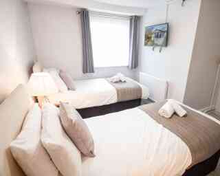 A cozy bedroom featuring two twin beds with white linens and gray accents. A lamp illuminates the room, and a flat-screen TV is mounted on the wall opposite the beds. Soft gray curtains frame the window.