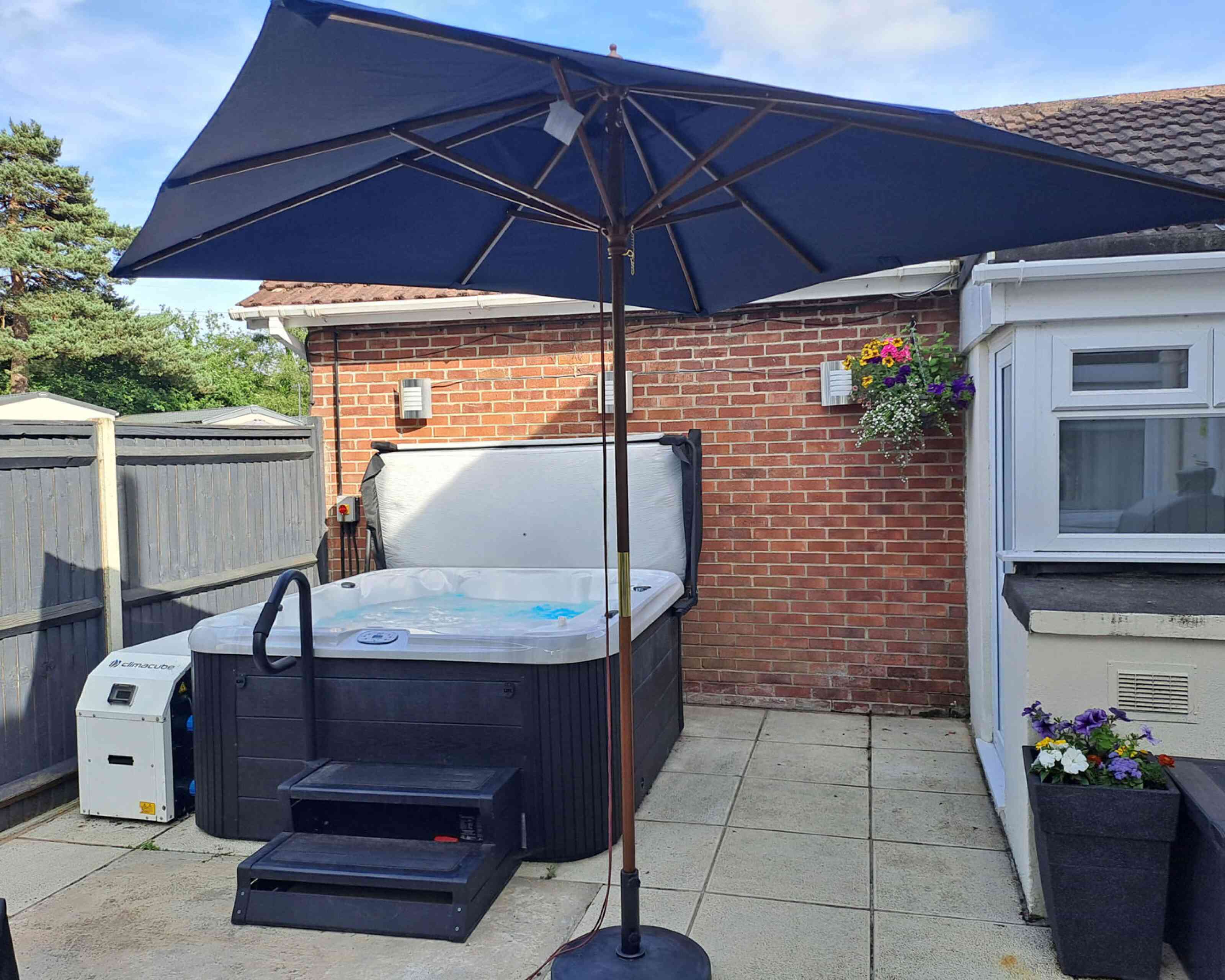 A hot tub with a raised lid is situated in a backyard. A large navy blue umbrella provides shade over the hot tub. Nearby, there is a plant hanging on the wall, and the area is paved with light-colored tiles.