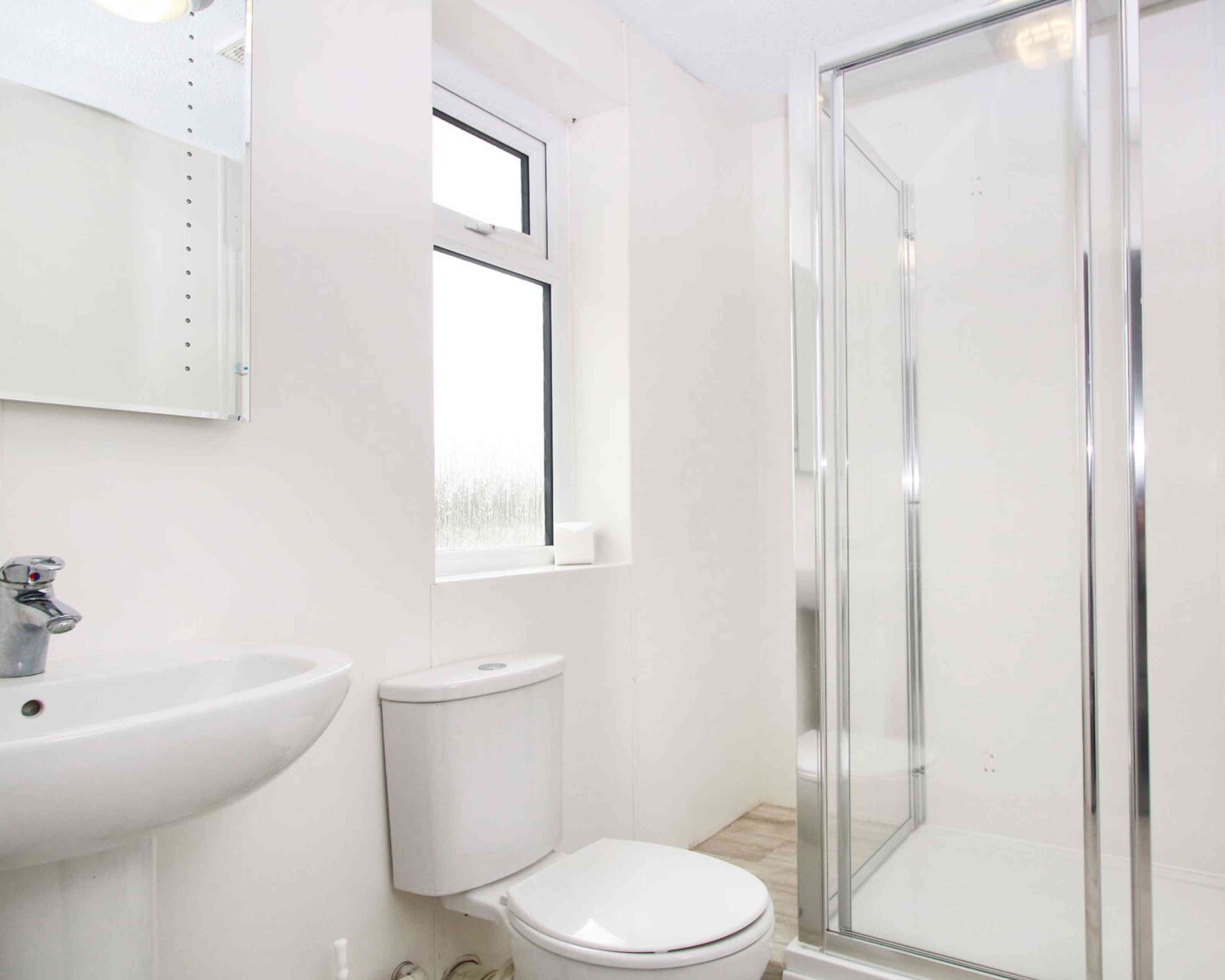 A modern bathroom featuring a shower stall with a glass door, a white sink, and a toilet. Natural light enters through a window. The walls are light-colored and the flooring is tiled.