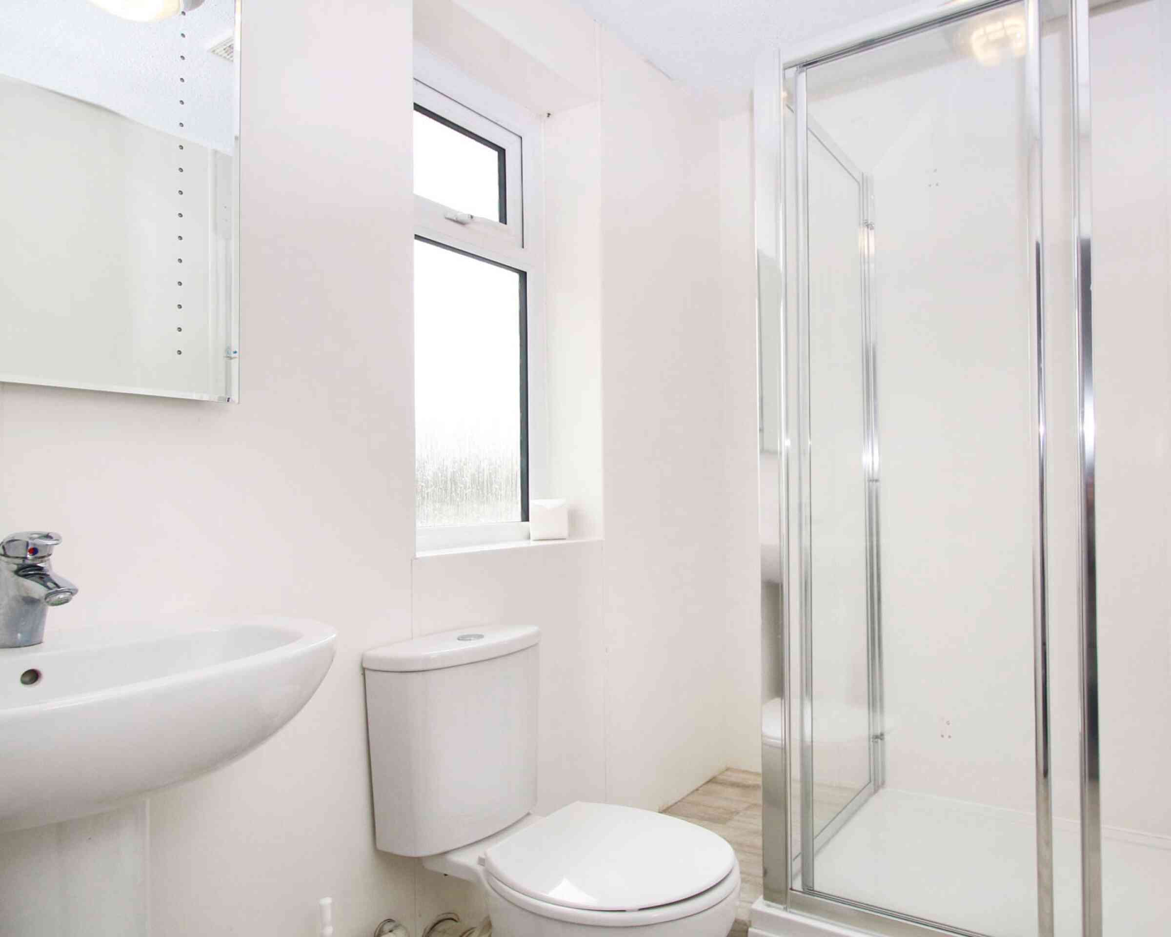 A modern bathroom featuring a shower stall with a glass door, a white sink, and a toilet. Natural light enters through a window. The walls are light-colored and the flooring is tiled.