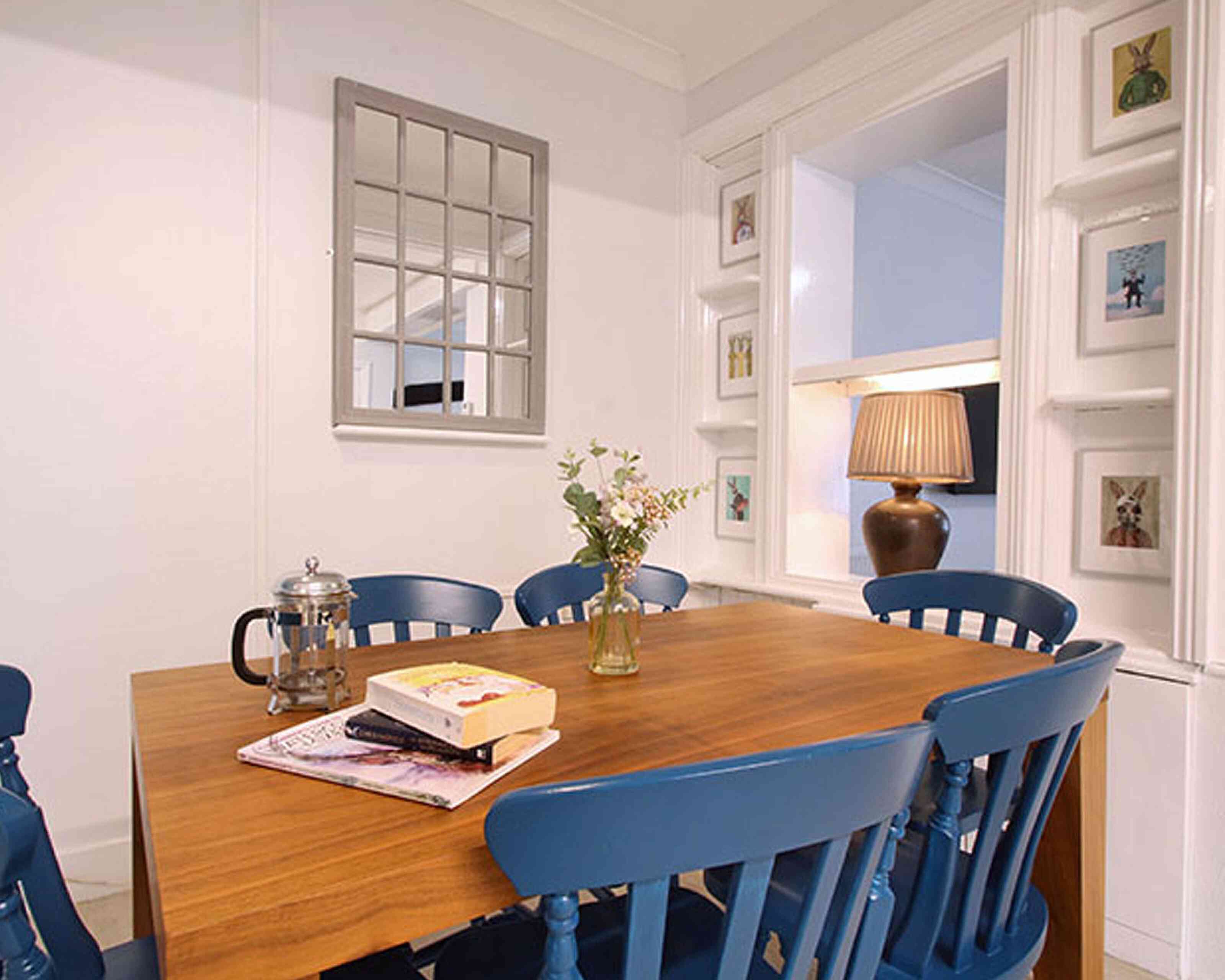 A cozy dining area features a wooden table surrounded by blue chairs. A small vase with flowers sits on the table, alongside a book and magazines. A mirror is mounted on the wall, and framed pictures are displayed on nearby shelves.