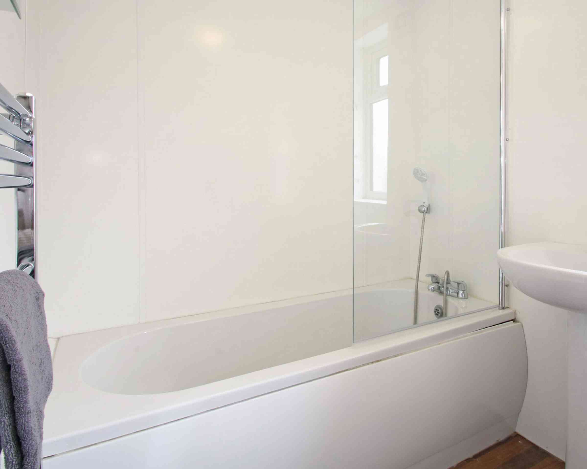 A modern bathroom featuring a white bathtub with a glass shower screen, a small sink, and a towel hanging on a rack. The walls are painted white, and the space is well-lit with natural light.