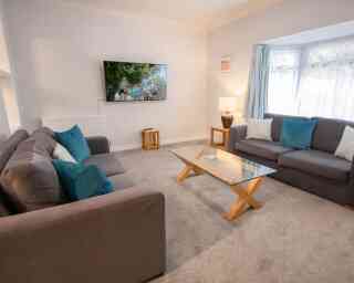 A cozy living room featuring two gray sofas with turquoise cushions, a glass coffee table with wooden legs, and a television mounted on the wall displaying a scenic outdoor scene. Natural light filters in through a window with sheer curtains.