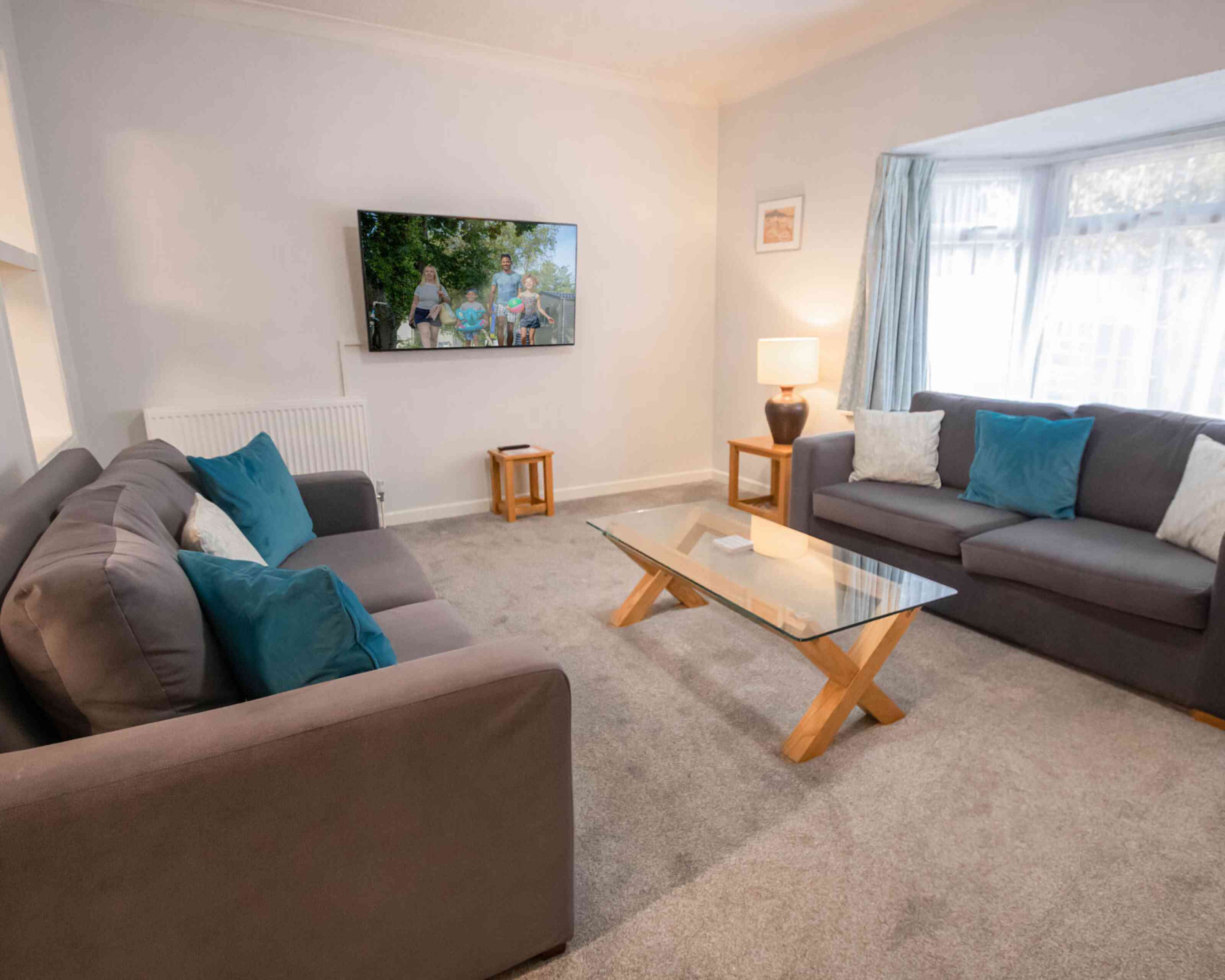 A cozy living room featuring two gray sofas with turquoise cushions, a glass coffee table with wooden legs, and a television mounted on the wall displaying a scenic outdoor scene. Natural light filters in through a window with sheer curtains.
