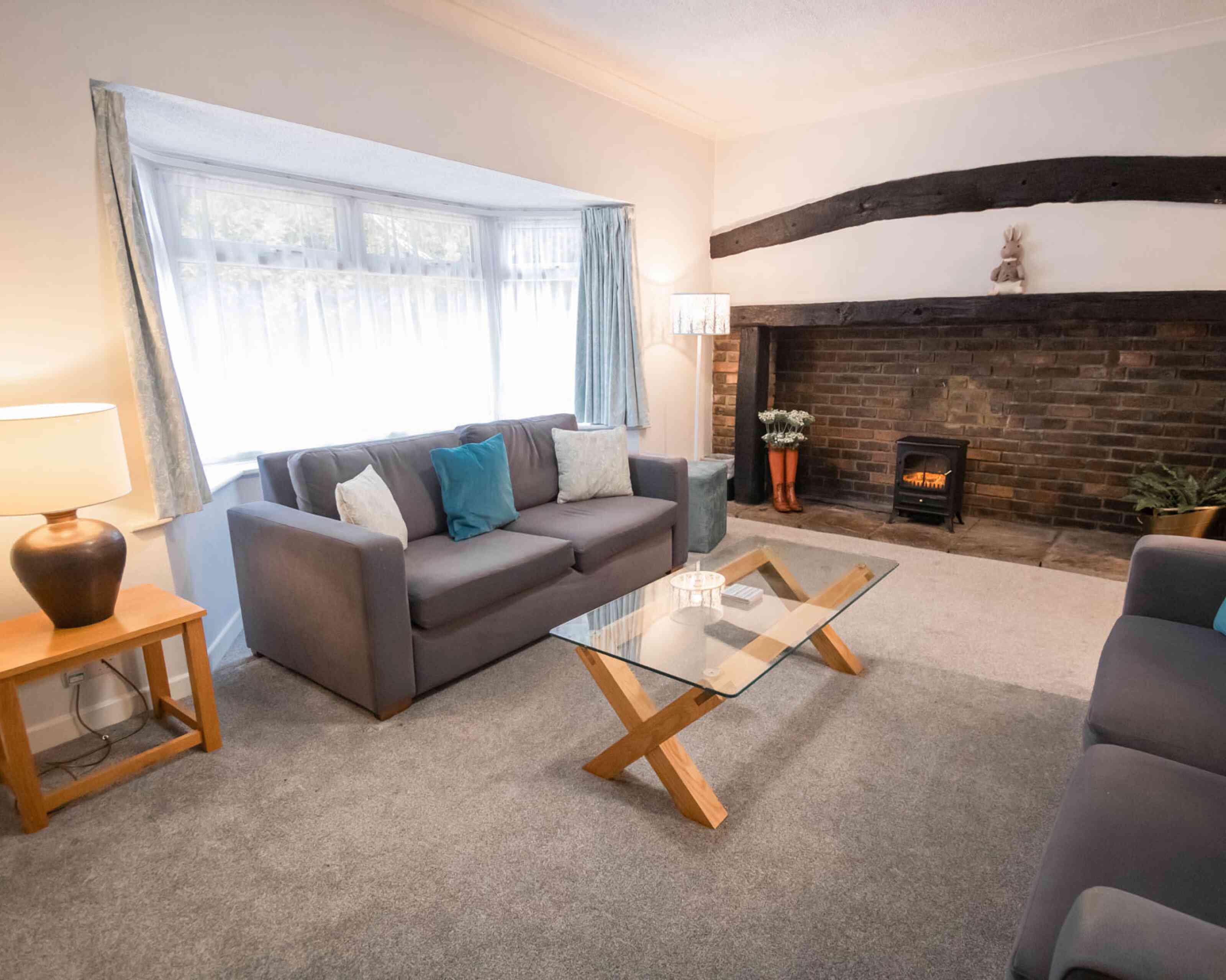 A cozy living room featuring a grey sofa with blue accent pillows, a glass coffee table with wooden legs, and a warm fireplace with a decorative mantle. Soft lighting from a lamp adds a welcoming ambiance. A large window allows natural light to fill the space.