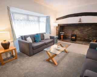 A cozy living room featuring a grey sofa with blue accent pillows, a glass coffee table with wooden legs, and a warm fireplace with a decorative mantle. Soft lighting from a lamp adds a welcoming ambiance. A large window allows natural light to fill the space.