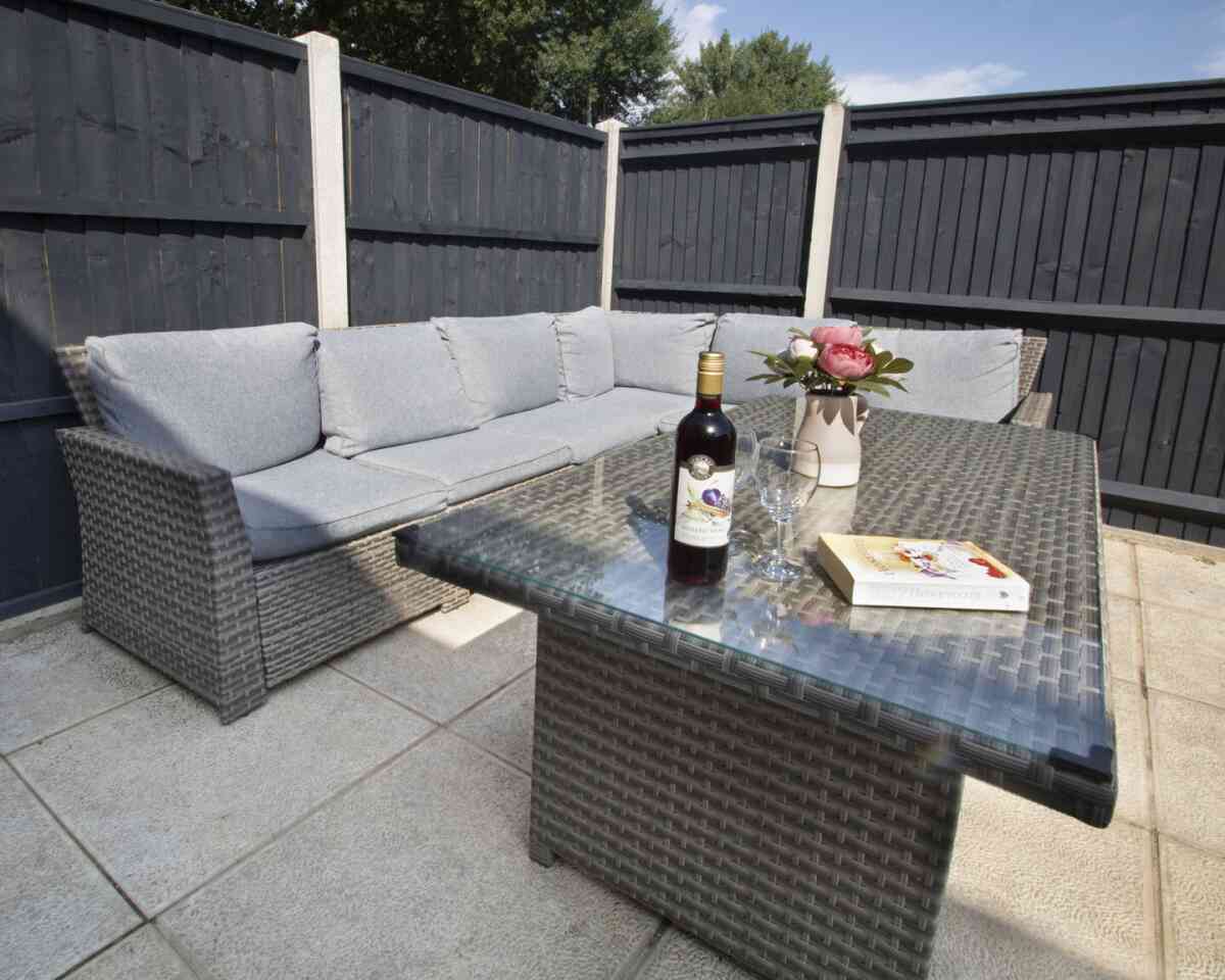 A cozy outdoor seating area featuring a gray sectional sofa with cushions, a glass-topped table, a bottle of wine, a glass, and a vase of pink roses, all set against dark wooden fences.
