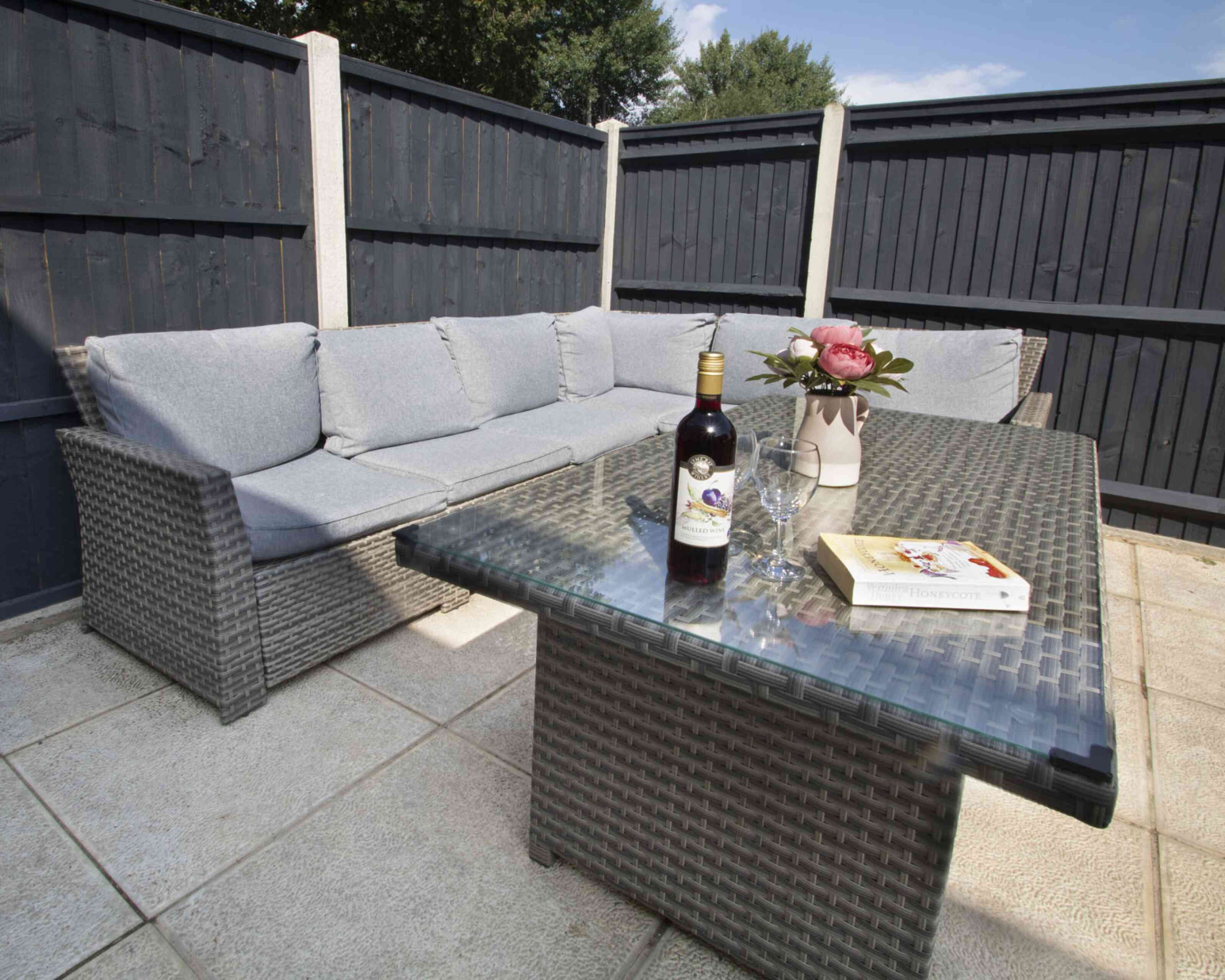 A cozy outdoor seating area featuring a gray sectional sofa with cushions, a glass-topped table, a bottle of wine, a glass, and a vase of pink roses, all set against dark wooden fences.