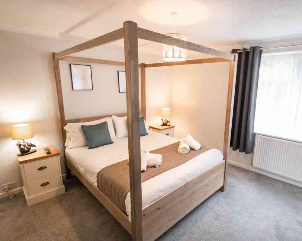 A cozy bedroom featuring a wooden four-poster bed with white bedding, two decorative pillows, and neatly folded towels. Soft lighting from two bedside lamps creates a warm ambiance, complemented by gray curtains and framed artwork on the walls.