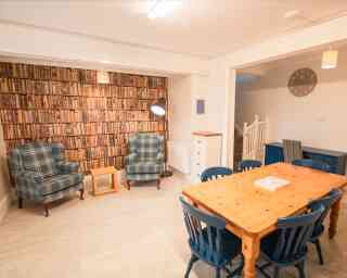 A cozy room featuring two plaid armchairs beside a small wooden table, a large wooden dining table with blue chairs, a decorative wall with a bookshelf design, and a clock on the wall.