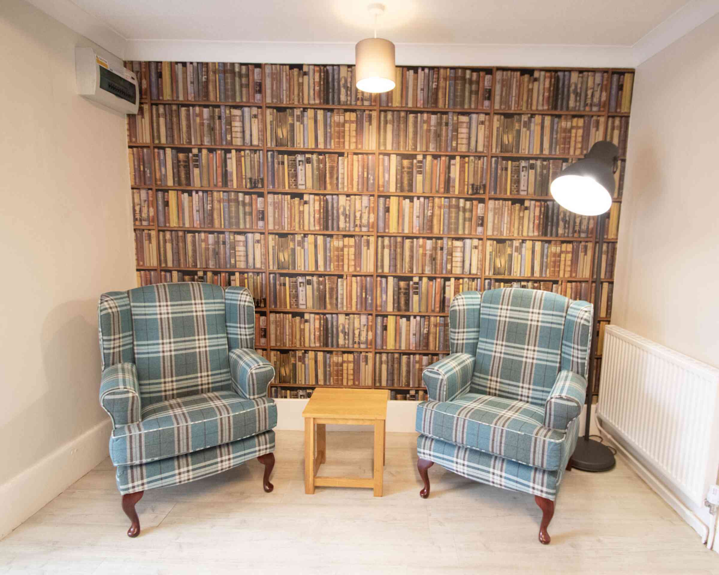A cozy corner with two plaid upholstered armchairs facing a small wooden table. The backdrop is a wall covered in a wallpaper design of stacked books, and a modern floor lamp stands nearby.