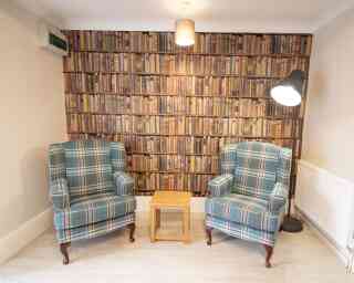 A cozy corner with two plaid upholstered armchairs facing a small wooden table. The backdrop is a wall covered in a wallpaper design of stacked books, and a modern floor lamp stands nearby.