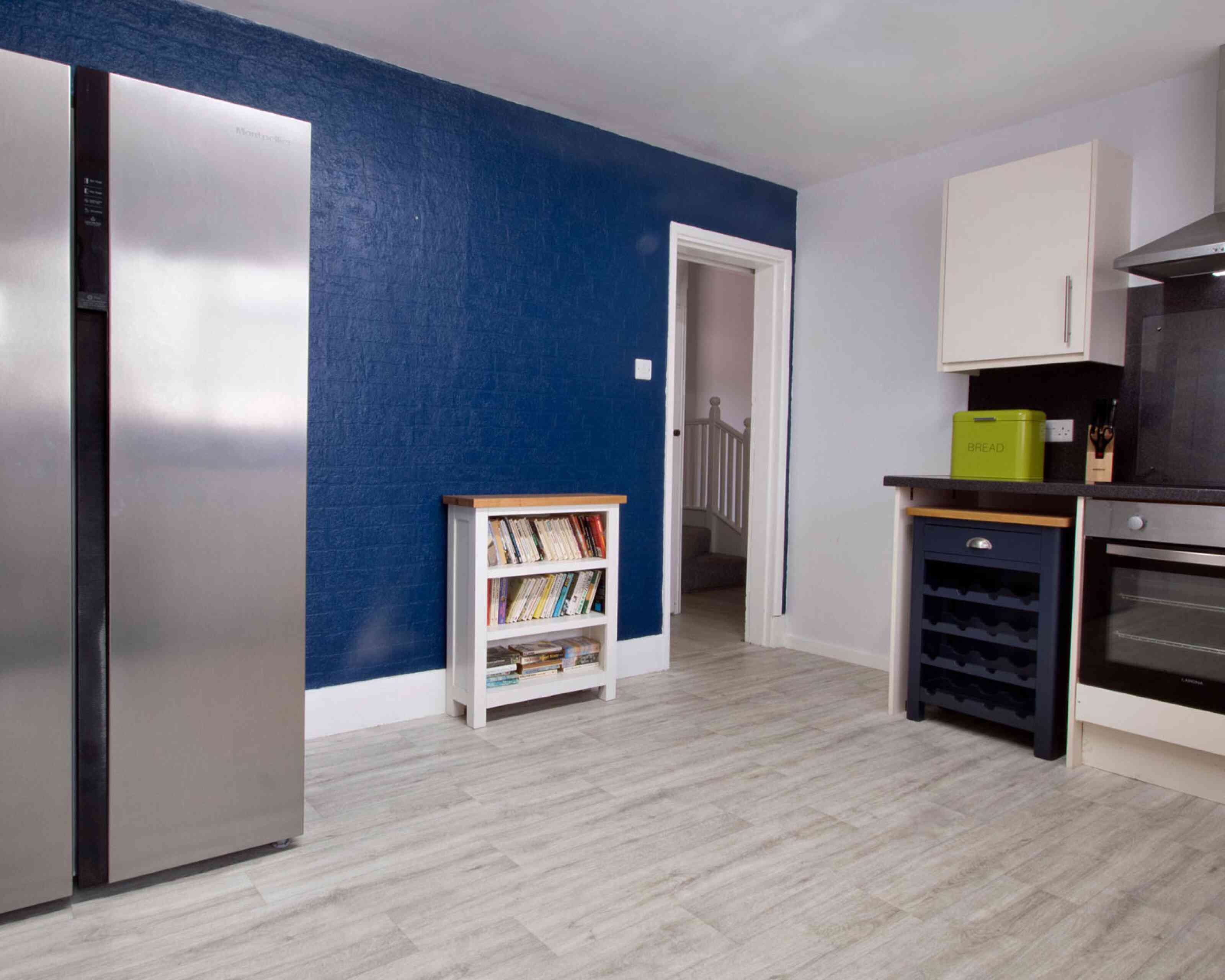 A modern kitchen featuring a stainless steel refrigerator, a white bookshelf with books, a dark countertop with a kitchen appliance, and a doorway leading to another room. The walls are painted blue and light gray.