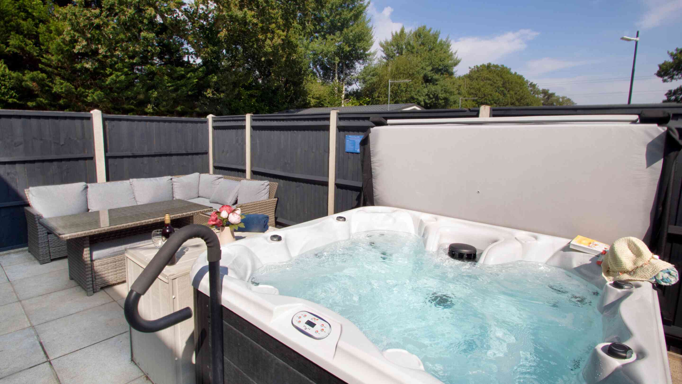 A private outdoor area featuring a hot tub with bubbling water, surrounded by a gray fence. Nearby, there's a seating area with a modern sofa and a small table, adorned with a bottle and flowers. Lush green trees provide a backdrop under a clear blue sky.