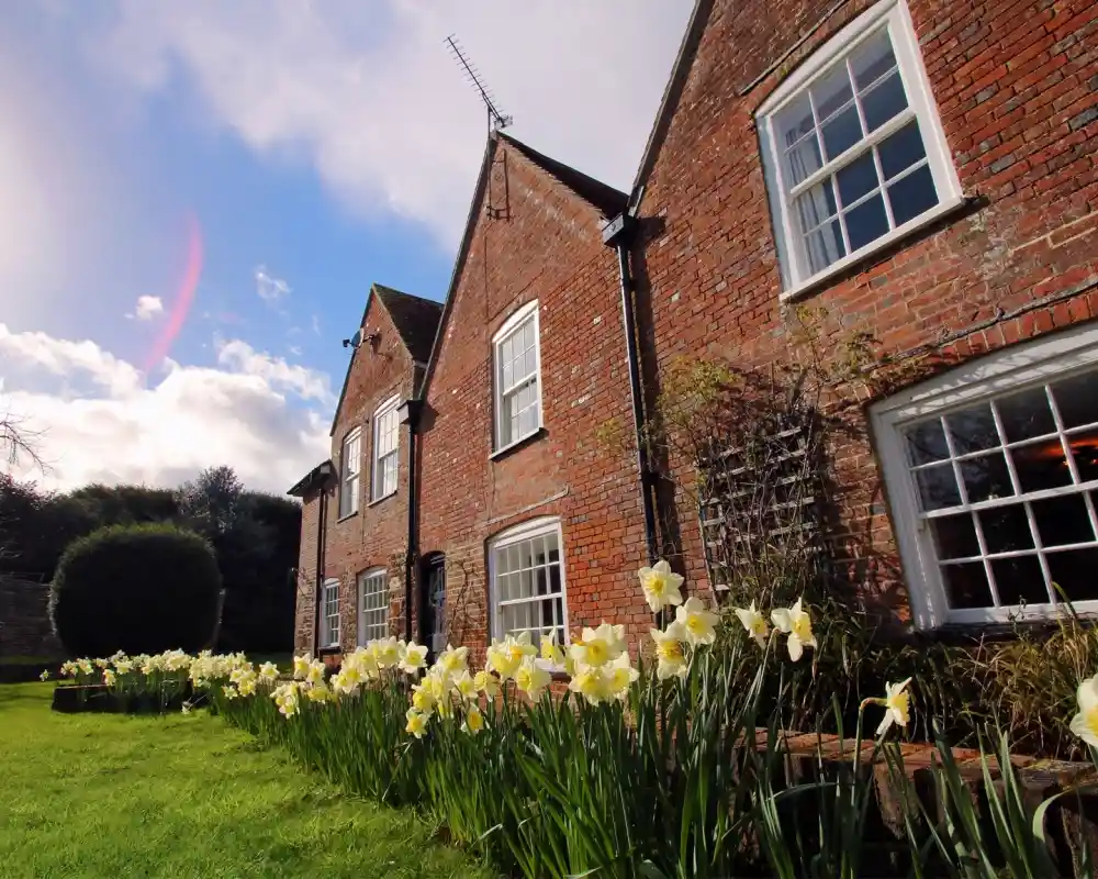 A charming red brick house with white windows is surrounded by blooming daffodils in a lush green garden. The sky is partly cloudy, adding a serene atmosphere to the scene.