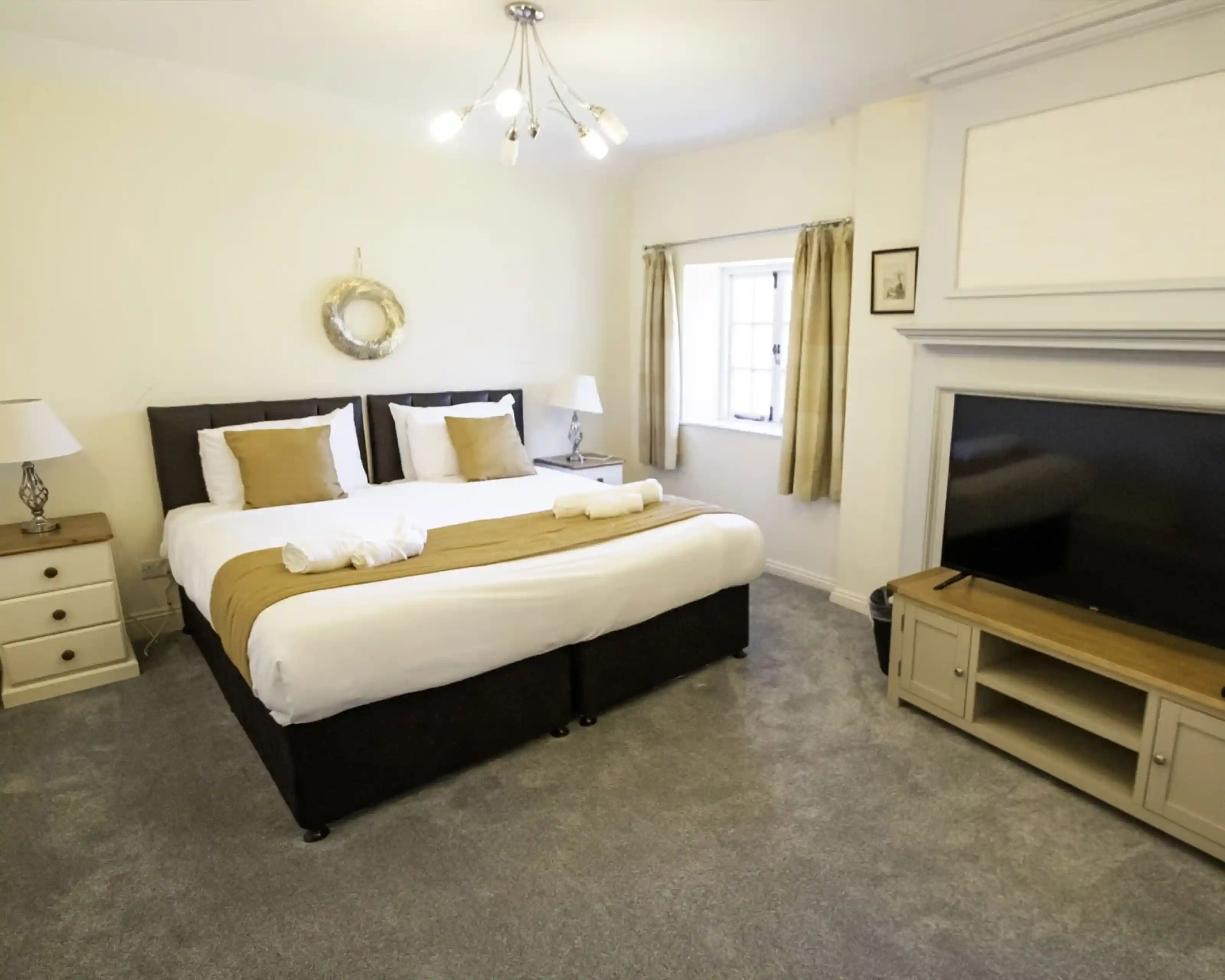 A cosy bedroom featuring a large bed with white and brown bedding, flanked by lamps on either side. There's a stylish dresser with drawers, a flat-screen TV on a wooden stand, and a window with beige curtains. The room has light-coloured walls and carpet.