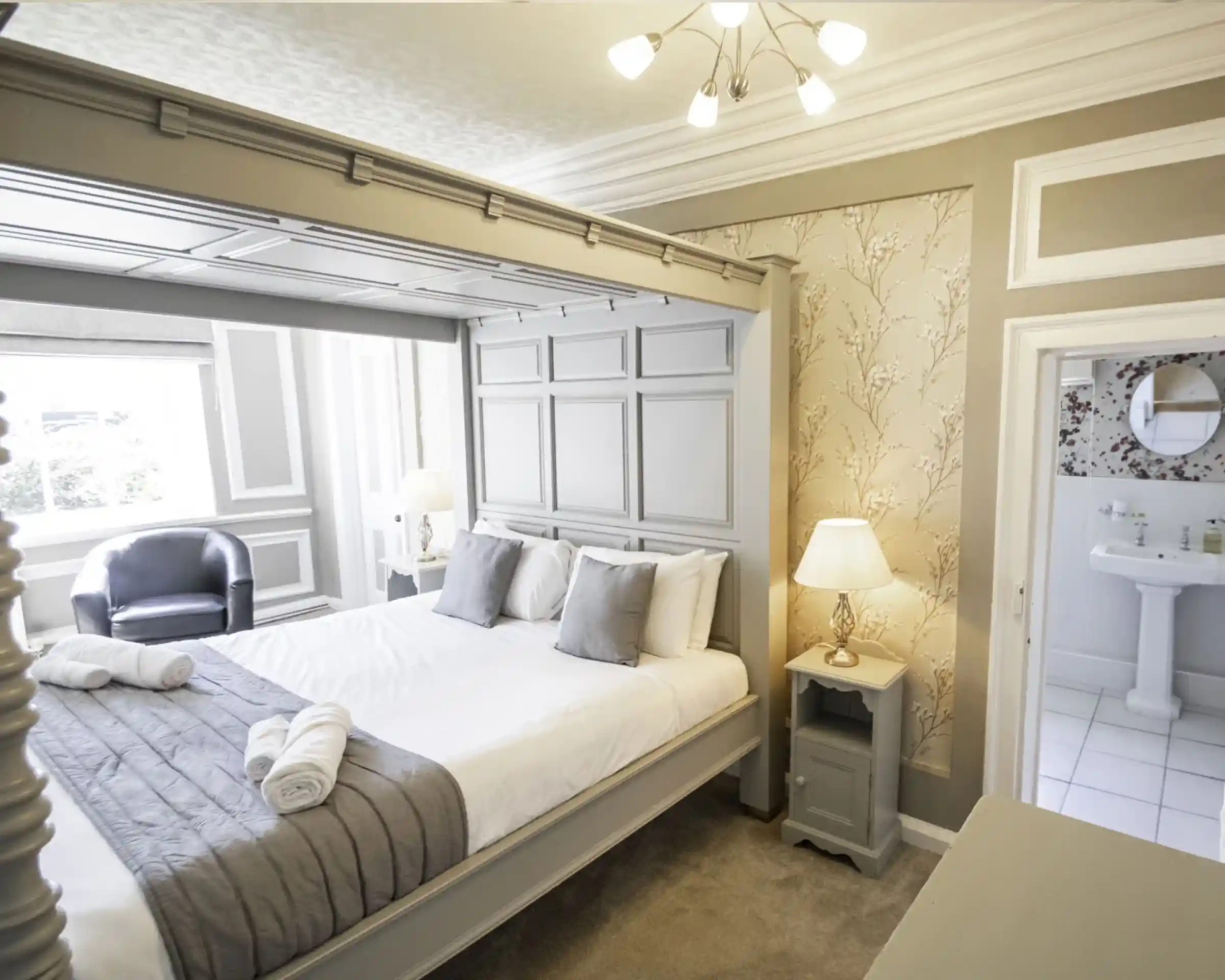 A cosy hotel room featuring a large grey bed with decorative headboard, two grey pillows, and neatly folded towels. A small bedside table with a lamp and a chair is visible, along with a bathroom area in the background. The walls are adorned with a subtle floral design.