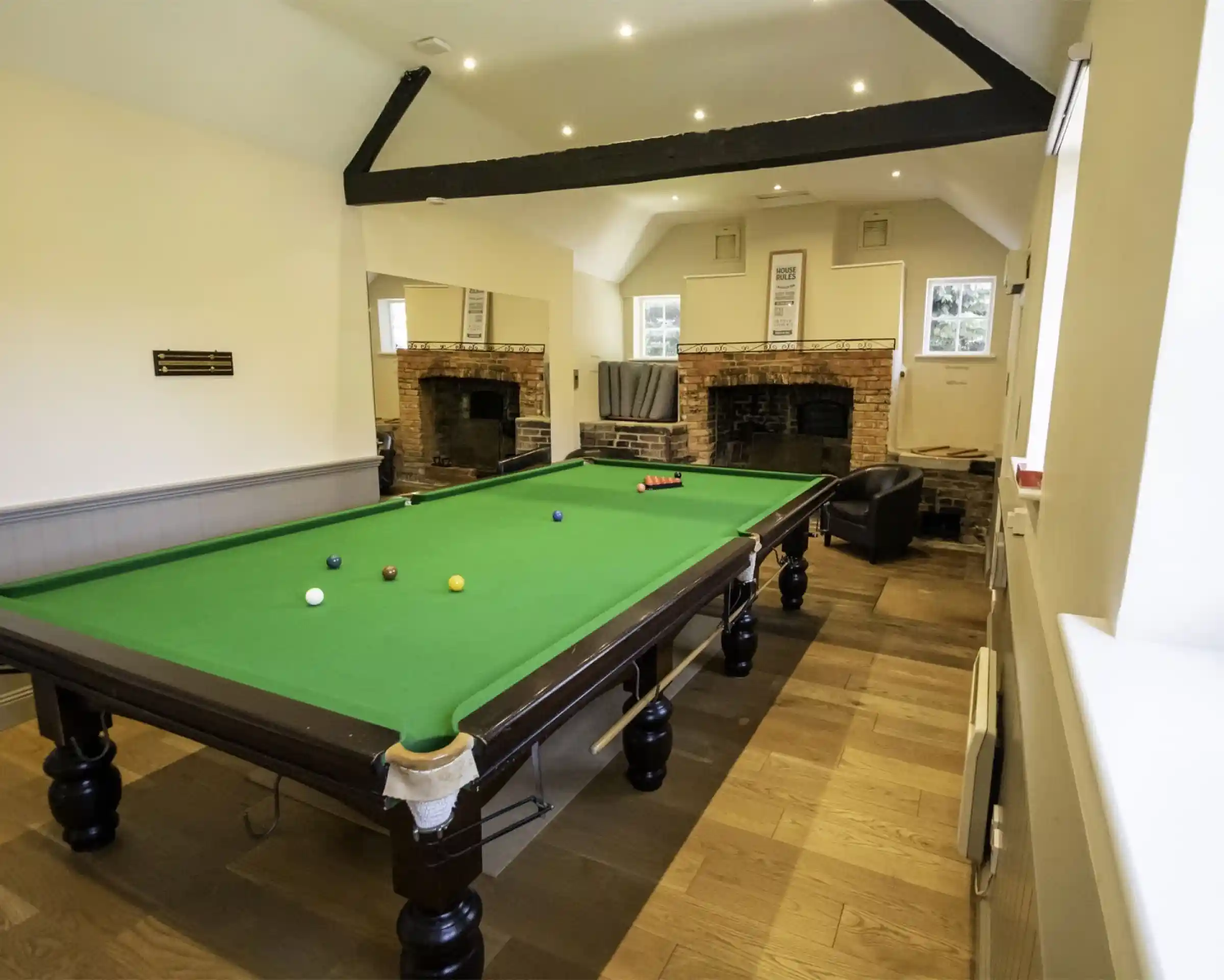 A spacious room featuring a green snooker table with balls arranged on it. The walls have a neutral colour, and there are two brick fireplaces. A large mirror reflects the room, and a comfortable chair is in the corner. Soft lighting complements the wooden flooring.