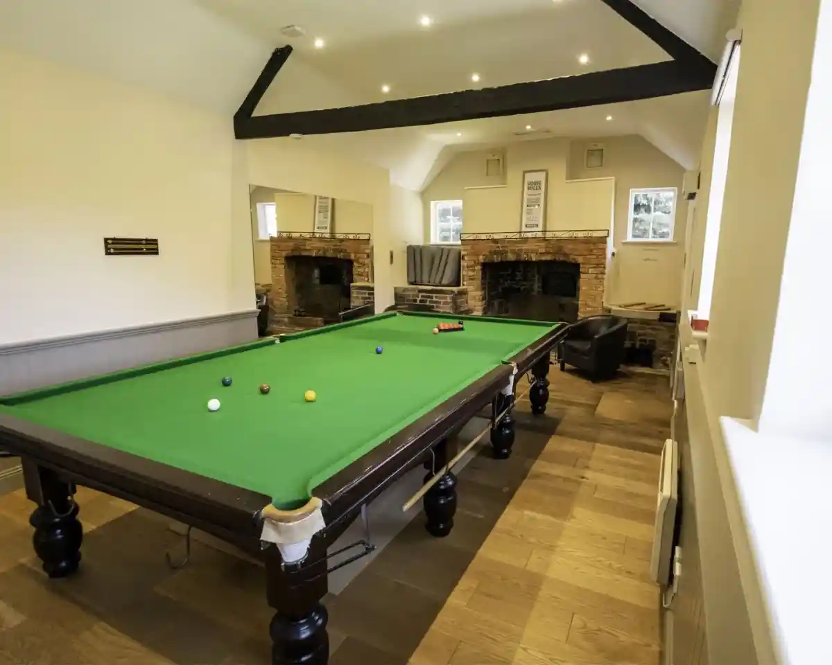 A spacious room featuring a green snooker table with balls arranged on it. The walls have a neutral colour, and there are two brick fireplaces. A large mirror reflects the room, and a comfortable chair is in the corner. Soft lighting complements the wooden flooring.
