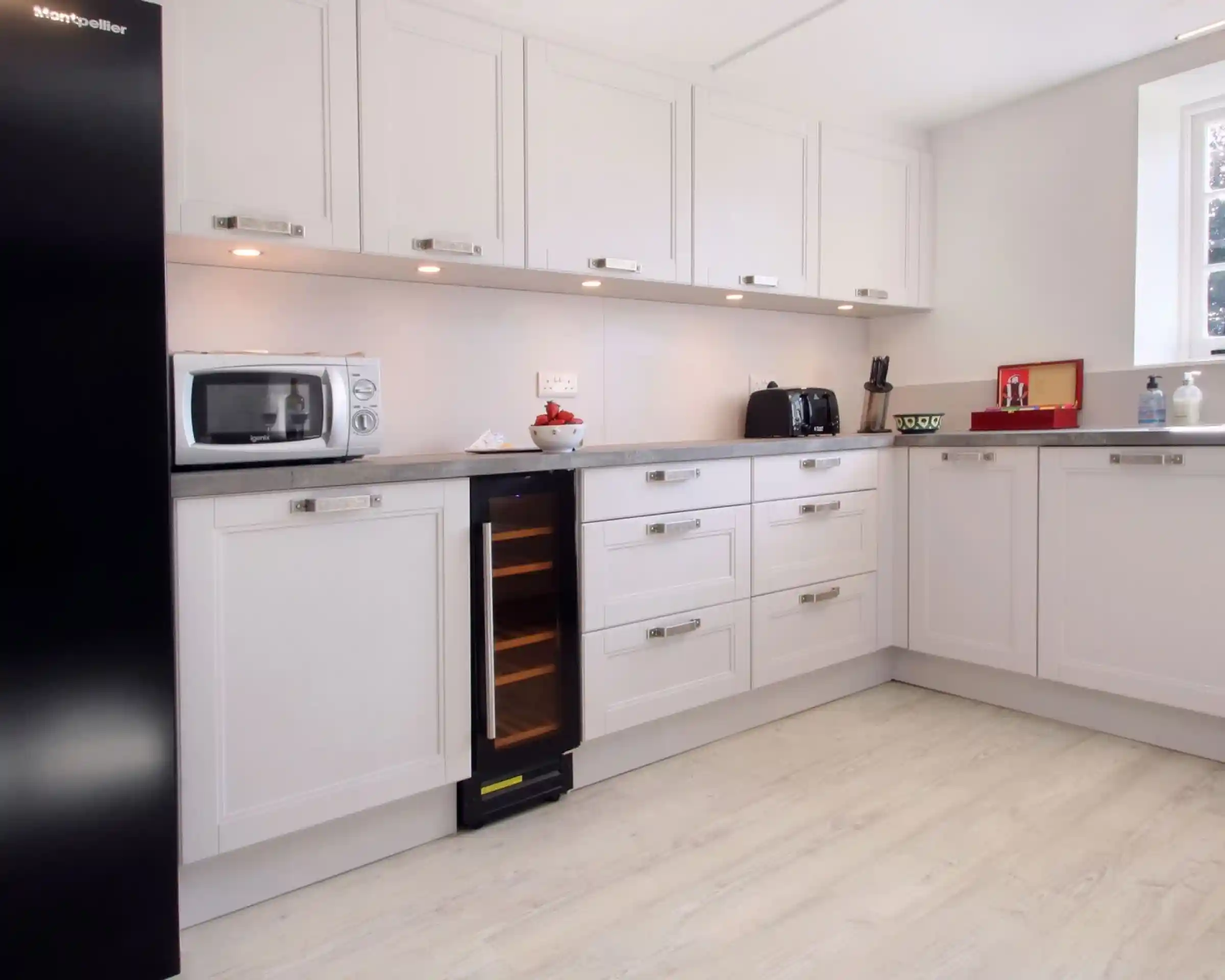 Modern kitchen featuring white cabinetry, a grey countertop, and stainless steel appliances including a microwave and a wine cooler. Bright and clean design with good lighting.