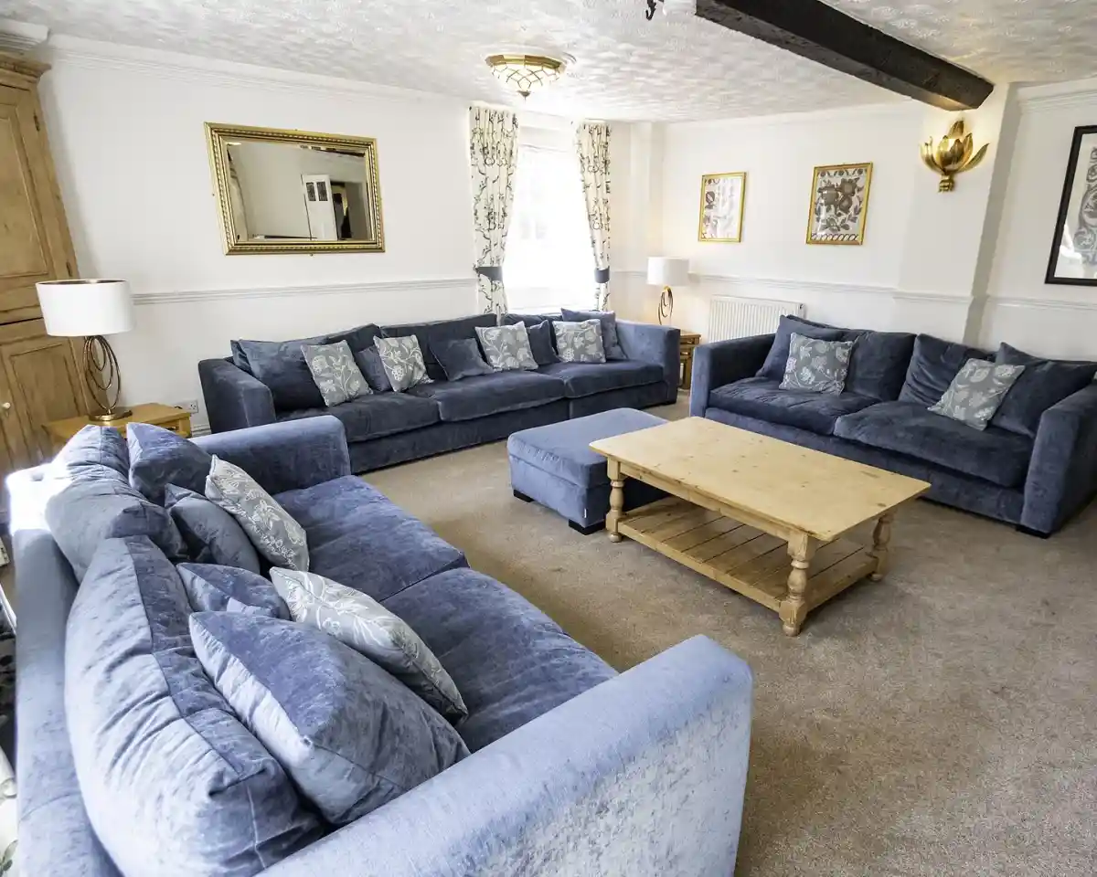 A spacious living room featuring two blue sofas arranged around a wooden coffee table. Decorative pillows adorn the sofas, and there is a large mirror and framed artwork on the walls. Elegant curtains and lighting fixtures enhance the inviting atmosphere.