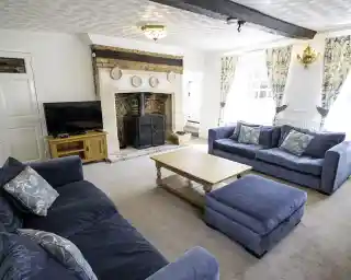 A spacious living room featuring two blue sofas, a coffee table, and an ottoman. A large fireplace with a decorative mantelpiece is at one end, and there is a television on a wooden stand. Natural light enters through large windows adorned with patterned curtains.