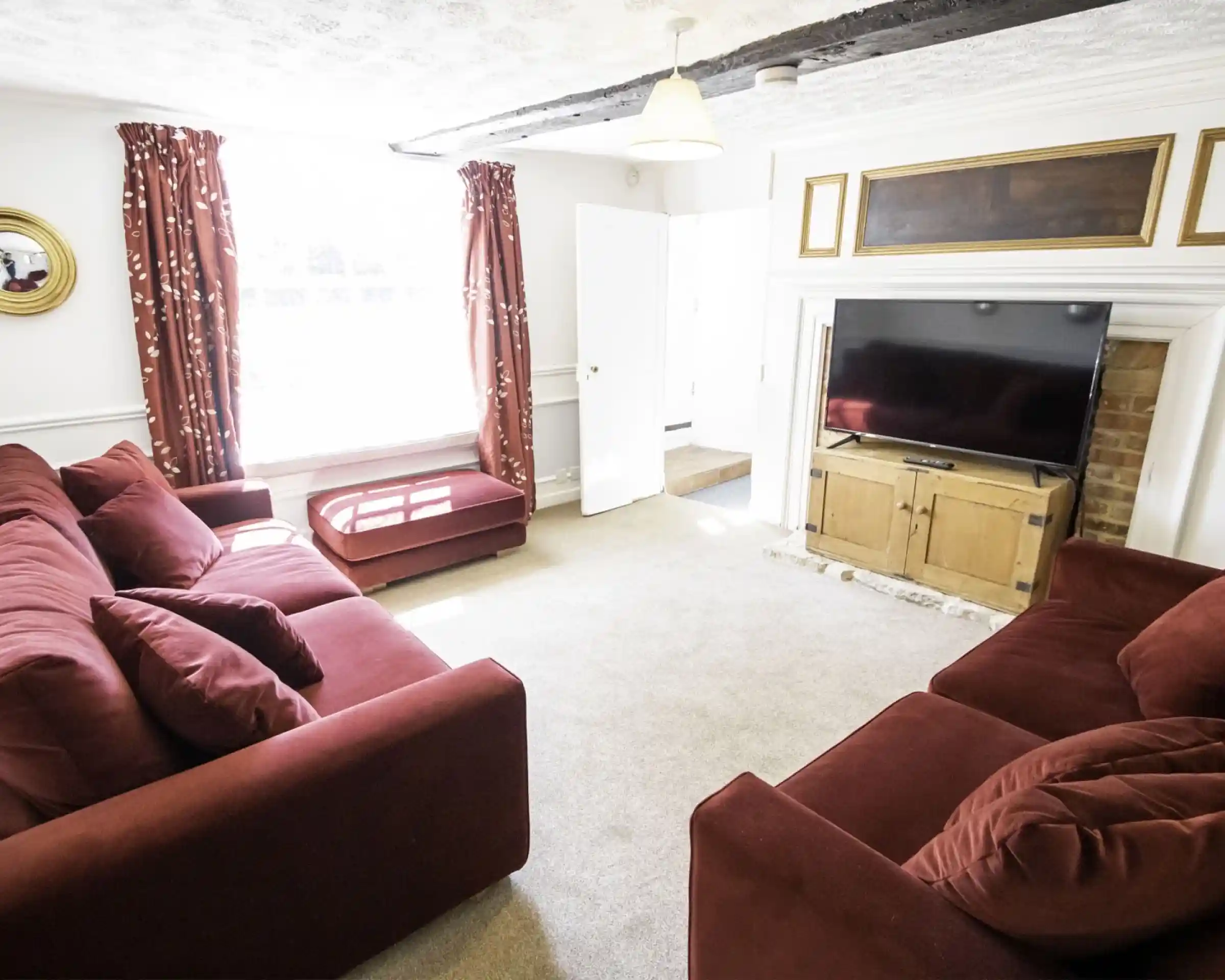 Cosy living room featuring two red sofas, a television on a wooden stand, and large windows with flowing curtains. A staircase is visible in the background, and the floor is carpeted.