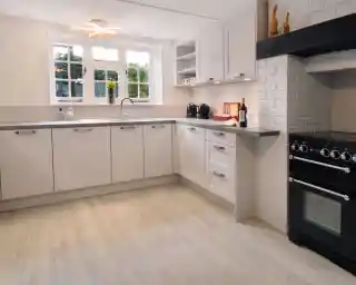 A modern kitchen featuring light cabinetry, a silver sink, and a black oven on the right. There is a countertop with small kitchen appliances and a ceiling fan above. Natural light enters through multiple windows, illuminating the space.