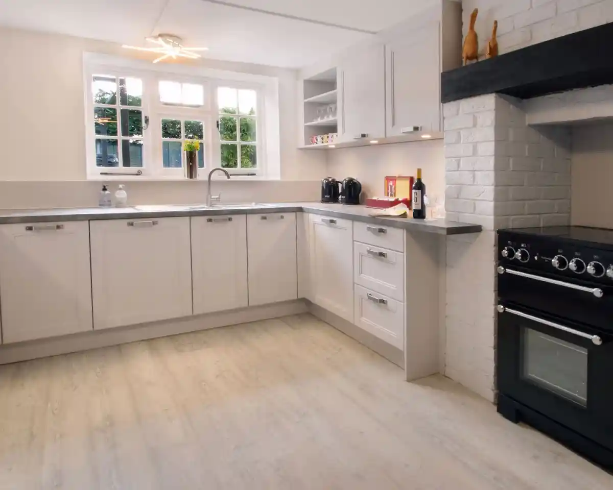 A modern kitchen featuring light cabinetry, a silver sink, and a black oven on the right. There is a countertop with small kitchen appliances and a ceiling fan above. Natural light enters through multiple windows, illuminating the space.