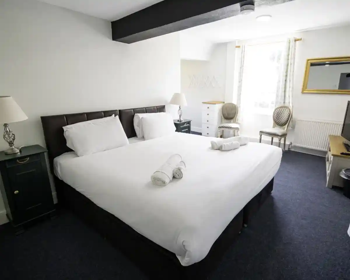 A spacious hotel room features a large bed with white linens, two rolled towels on the bed, bedside tables with lamps, a dresser, and two elegant chairs by a window. A mirror hangs on the wall, adding to the bright and inviting atmosphere.