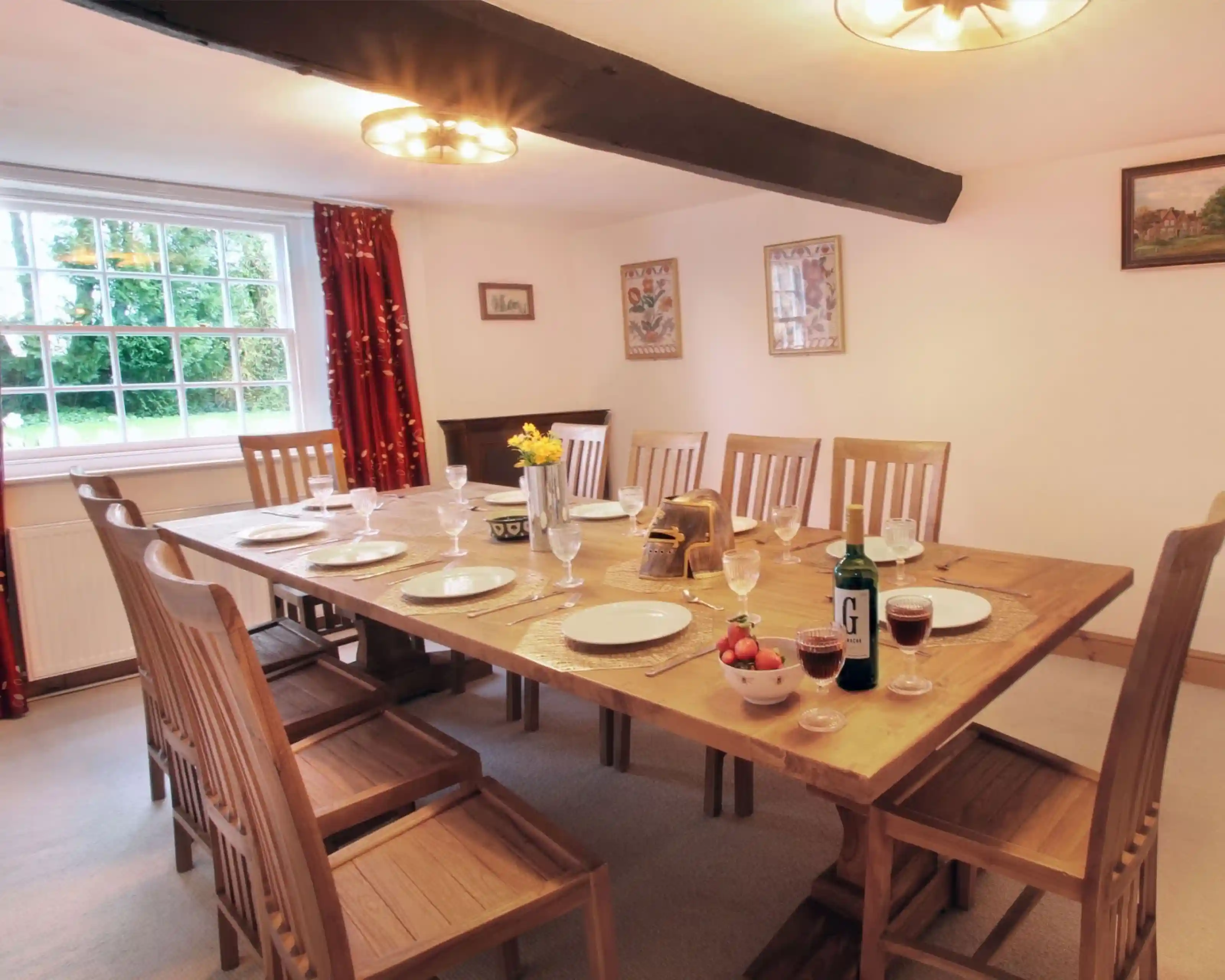A spacious dining room features a large wooden table set for a meal with plates, glasses, and a centrepiece of flowers. Twelve wooden chairs surround the table. Warm lighting and red curtains add a cosy atmosphere. A window allows natural light to brighten the space.