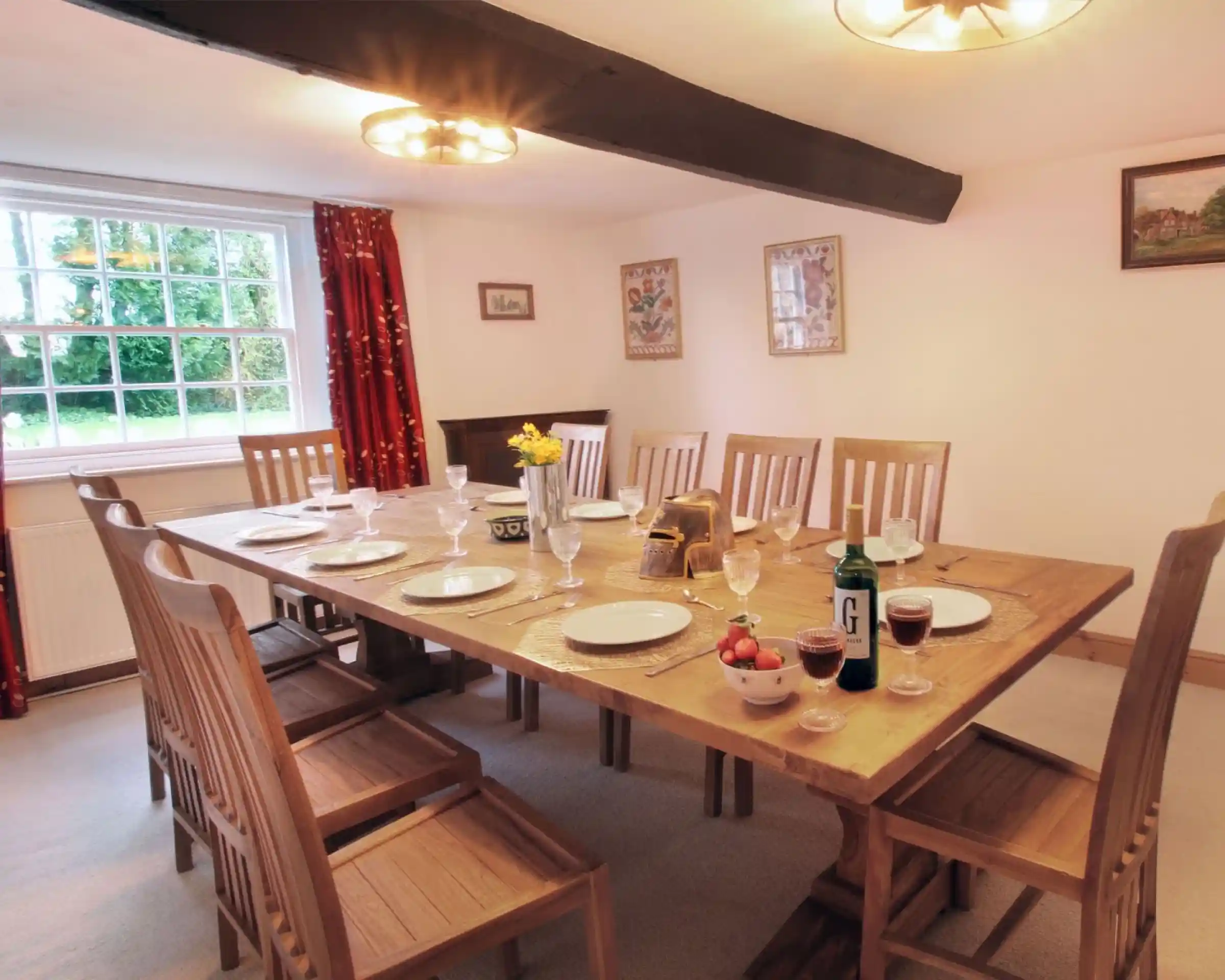 A spacious dining room features a large wooden table set for a meal with plates, glasses, and a centrepiece of flowers. Twelve wooden chairs surround the table. Warm lighting and red curtains add a cosy atmosphere. A window allows natural light to brighten the space.