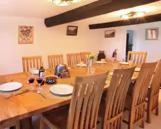 A spacious dining area featuring a large wooden table set with plates, glasses, and a bottle of wine. Eight wooden chairs are arranged around the table, with decorative accents on the walls and a cozy atmosphere highlighted by ceiling lights.