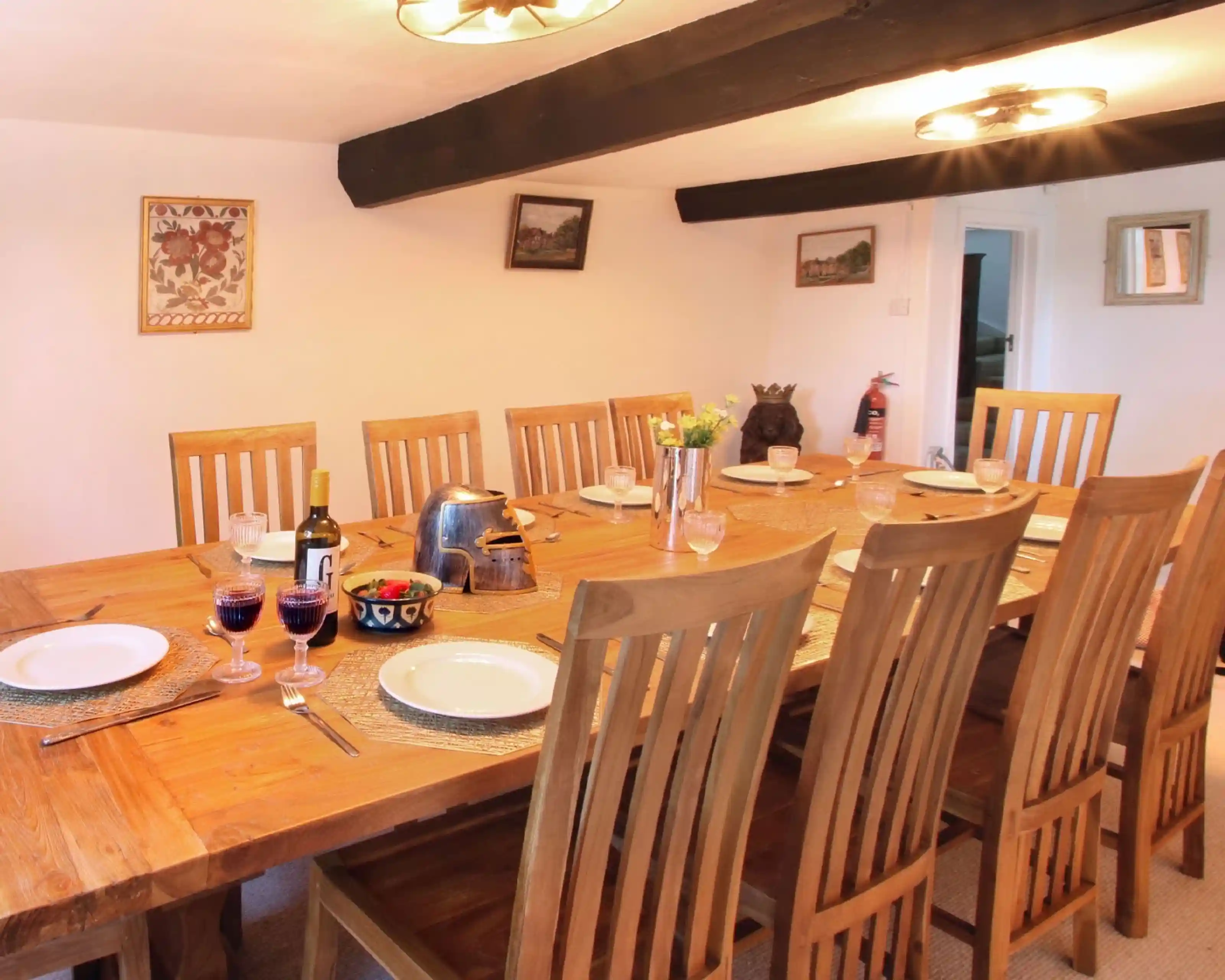 A spacious dining area featuring a large wooden table set with plates, glasses, and a bottle of wine. Eight wooden chairs are arranged around the table, with decorative accents on the walls and a cozy atmosphere highlighted by ceiling lights.